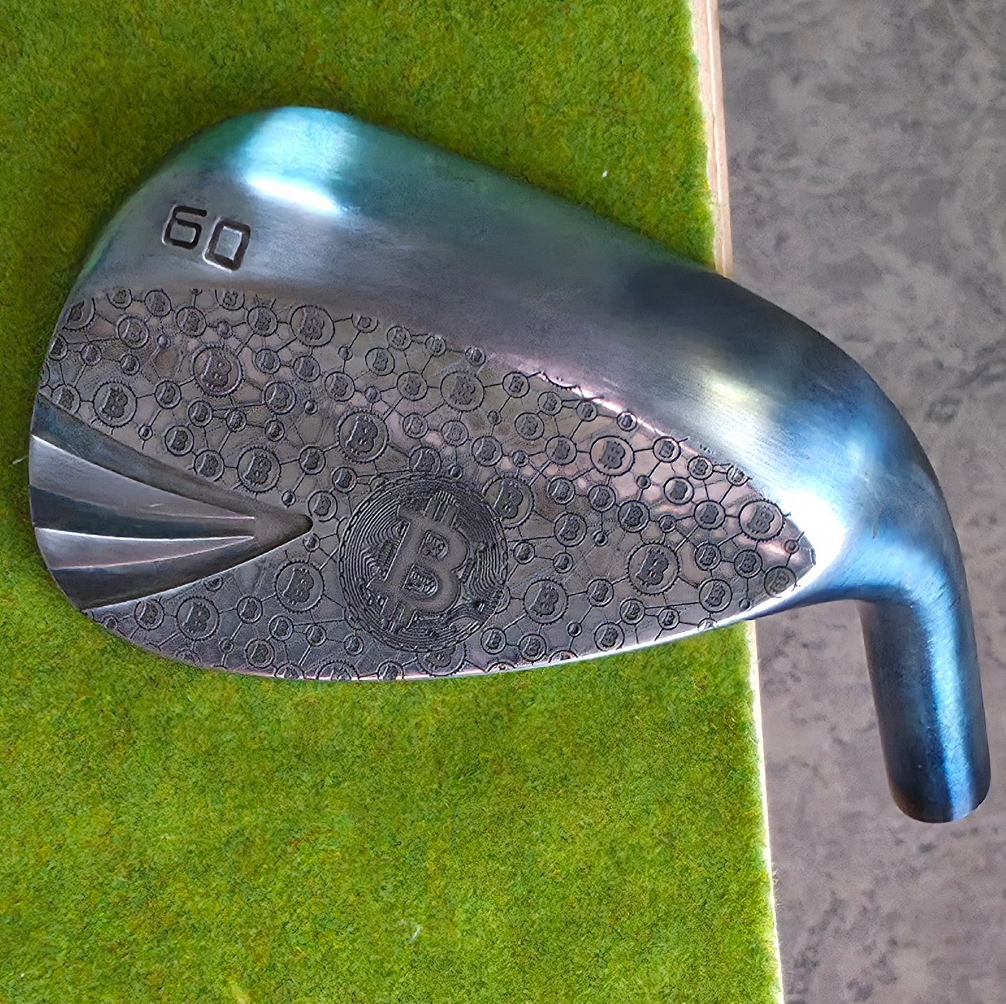 A 60-degree golf lob wedge featuring a custom laser-engraved Bitcoin theme. The back of the metal clubhead displays a central Bitcoin logo surrounded by a complex network of smaller Bitcoin icons and interconnected blockchain nodes. The club face features standard scoring grooves with a detailed engraving of a Bitcoin rocket ship on the toe. Resting on vibrant green artificial turf, this unique wedge seamlessly combines modern cryptocurrency culture with premium custom golf equipment design.