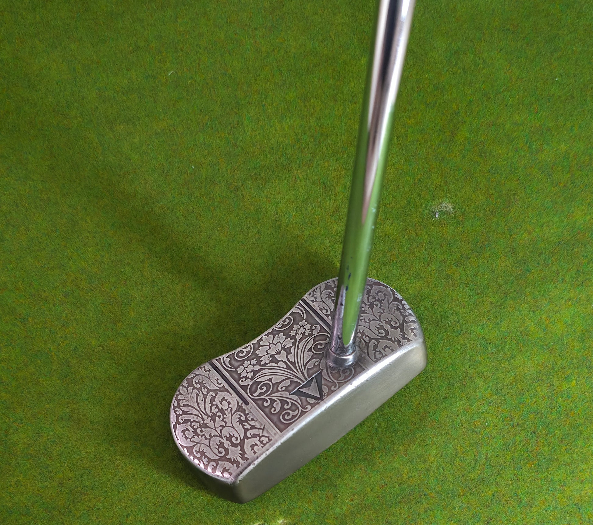 Golf putter with decorative head on a green background Custom engraved golf putter head with ornate floral and scrollwork design in antique brushed metal finish. Features deep engraving for durability, luxury artisan detailing, and an elegant symmetrical pattern. Perfect for golfers seeking a unique club, golf collectors, or as a personalized gift with initials, logos, or dates added. A blend of performance and craftsmanship designed to stand out on the green or in a golf display collection.