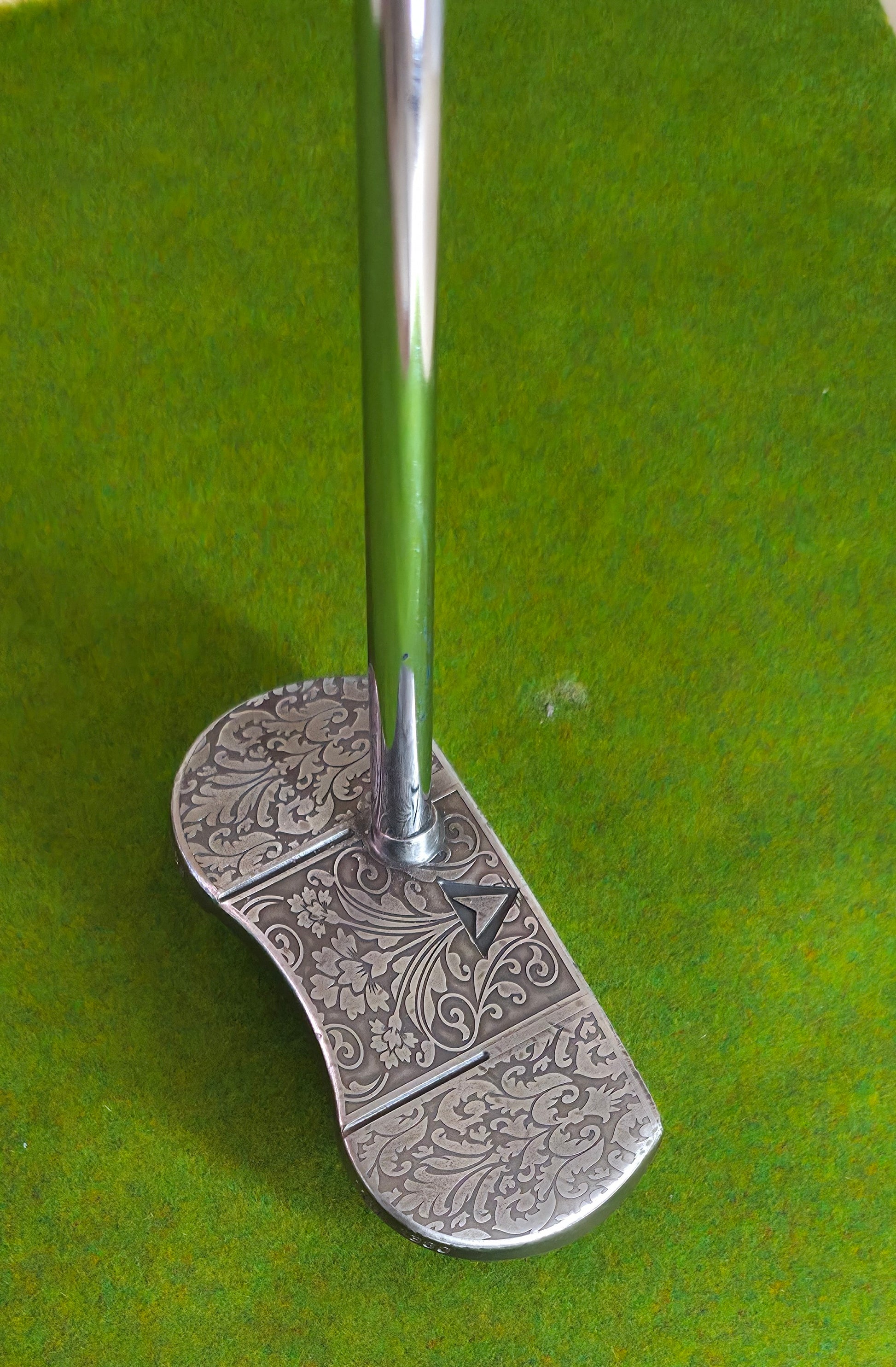 Golf club with decorative head on a green background Custom engraved golf putter head with ornate floral and scrollwork design in antique brushed metal finish. Features deep engraving for durability, luxury artisan detailing, and an elegant symmetrical pattern. Perfect for golfers seeking a unique club, golf collectors, or as a personalized gift with initials, logos, or dates added. A blend of performance and craftsmanship designed to stand out on the green or in a golf display collection.