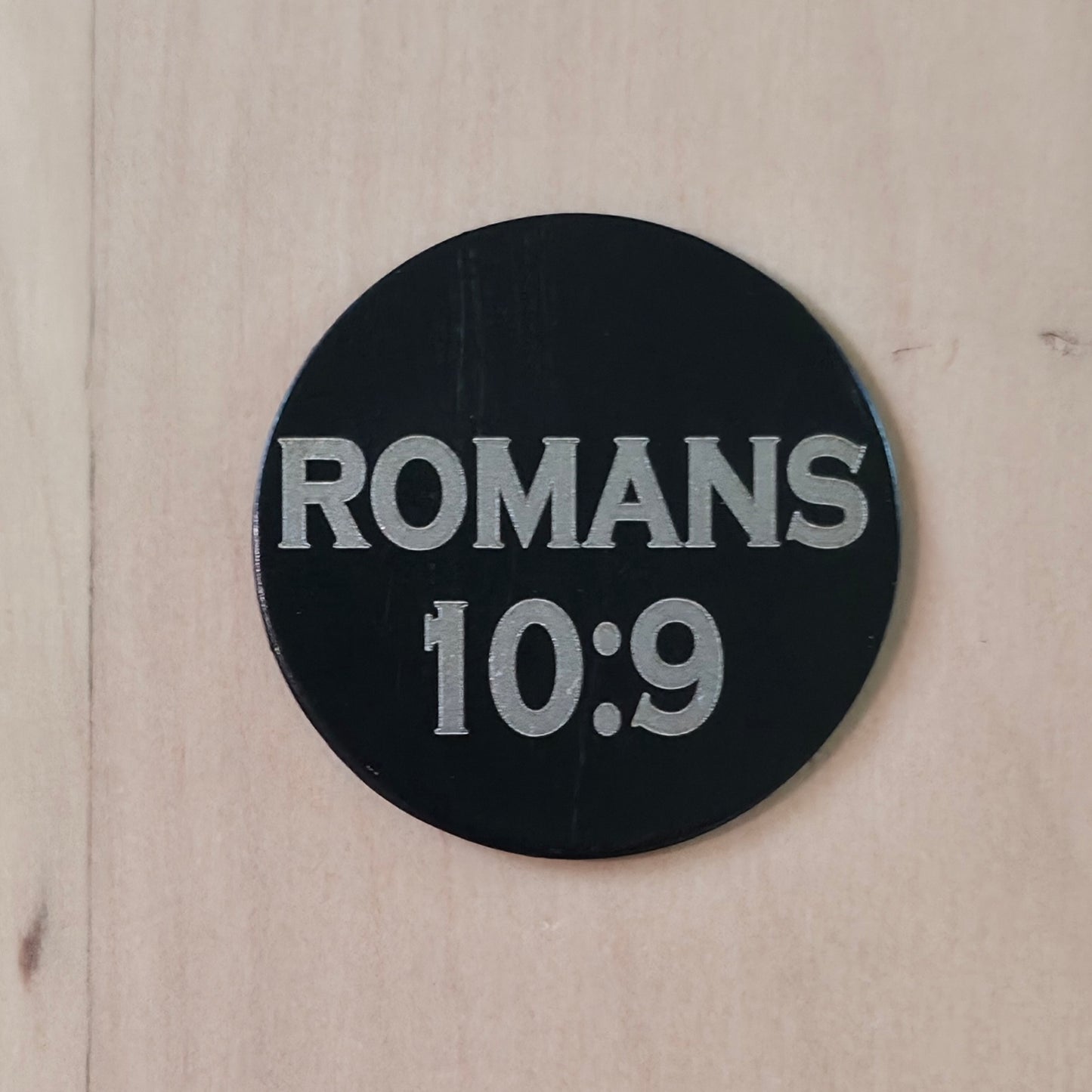 A round black sticker or coaster with the text 'ROMANS 10:9' in light gray, shown on a light wood background.