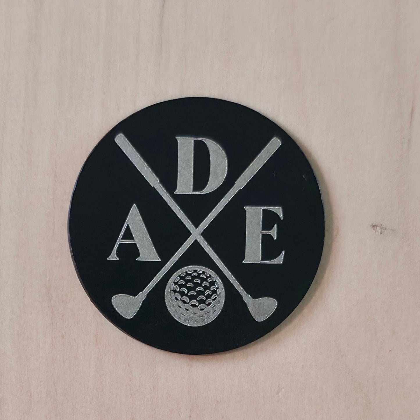A round black sticker or patch featuring the letters D, A, and E in white, crossed with golf clubs and a golf ball, displayed on a light wood background.
