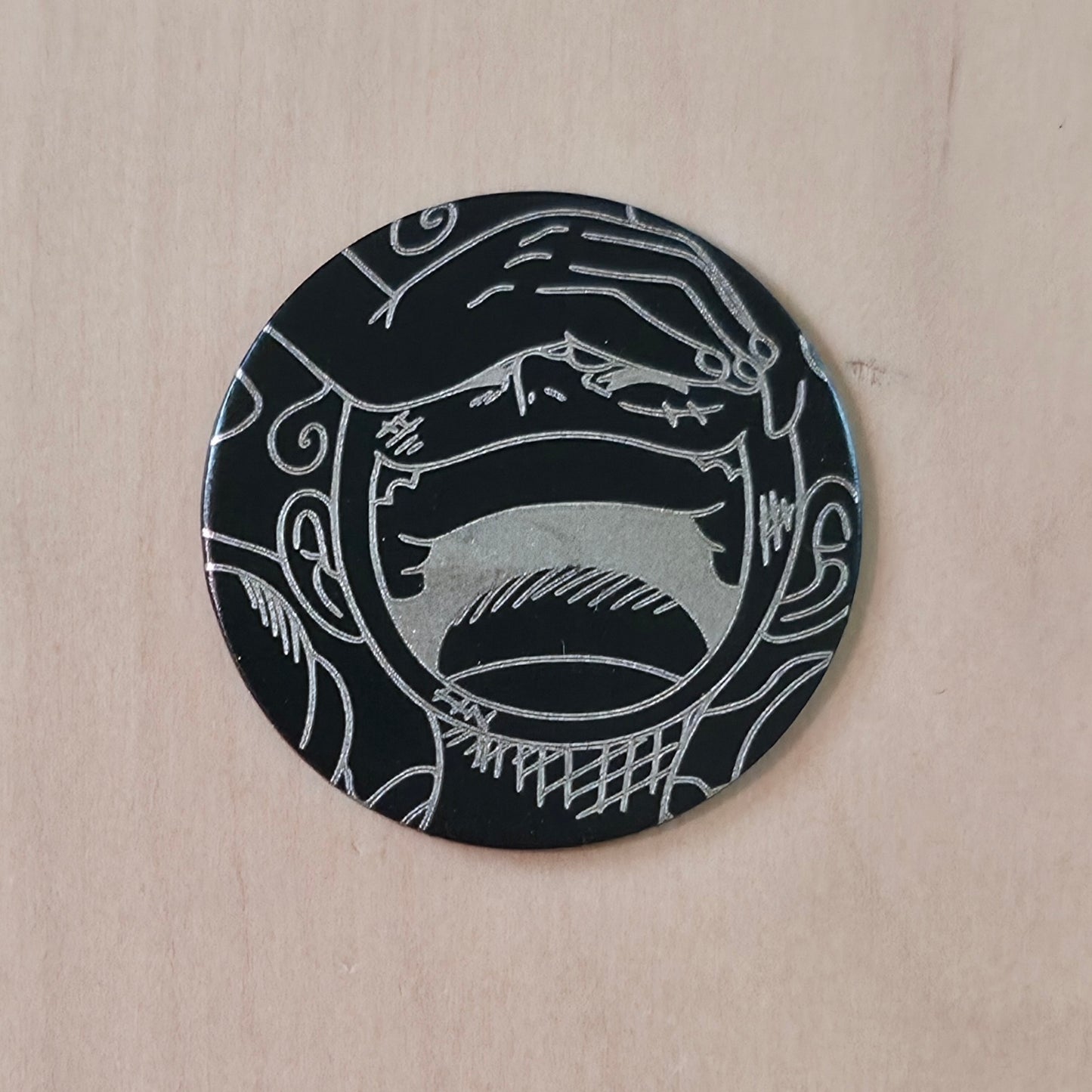 A round black coaster with a white engraved graphic of a screaming face, displayed on a light wood background.