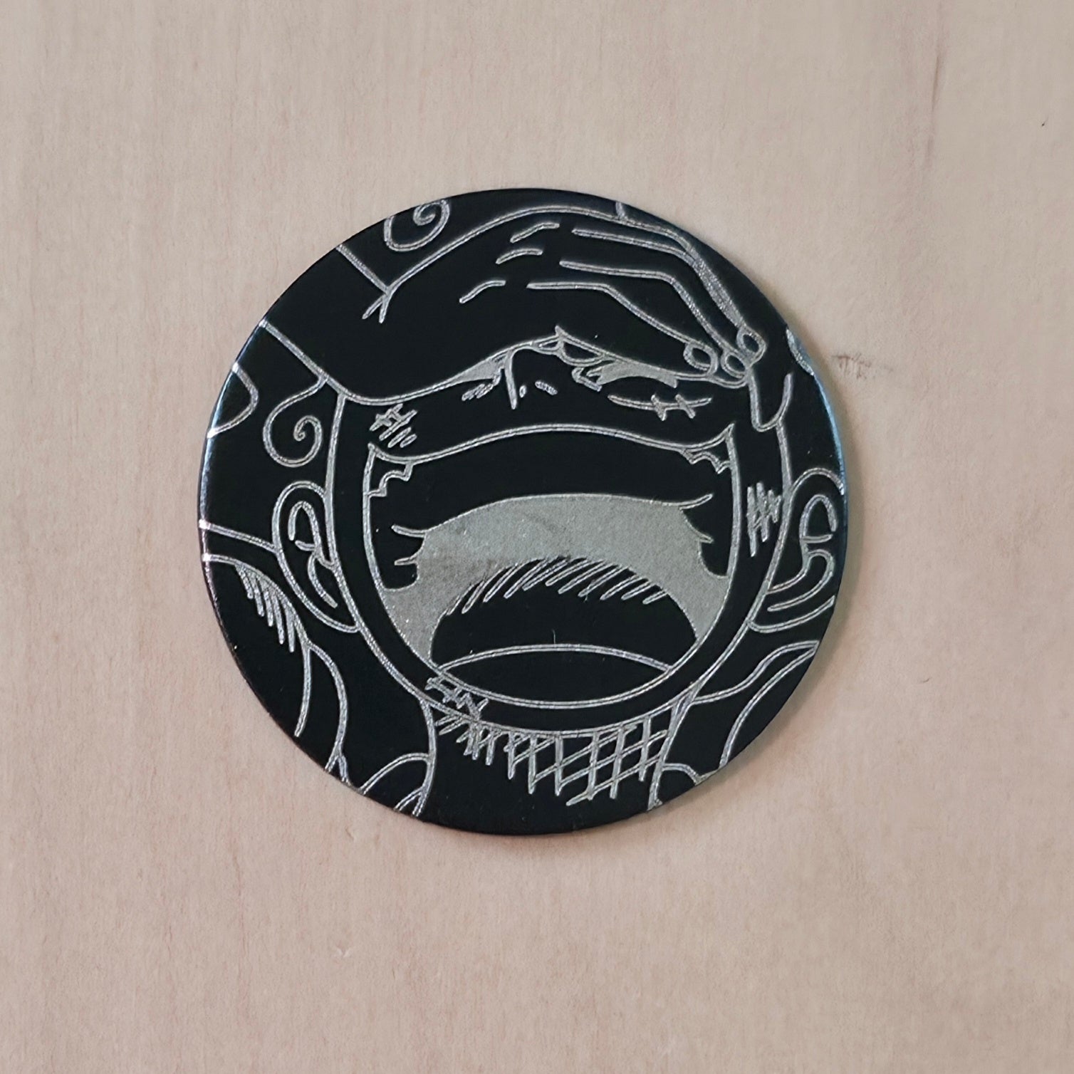 A round black coaster with a white engraved graphic of a screaming face, displayed on a light wood background.