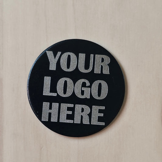 A round black custom badge or sticker with silver glitter text reading 'YOUR LOGO HERE', displayed on a light beige wood background.