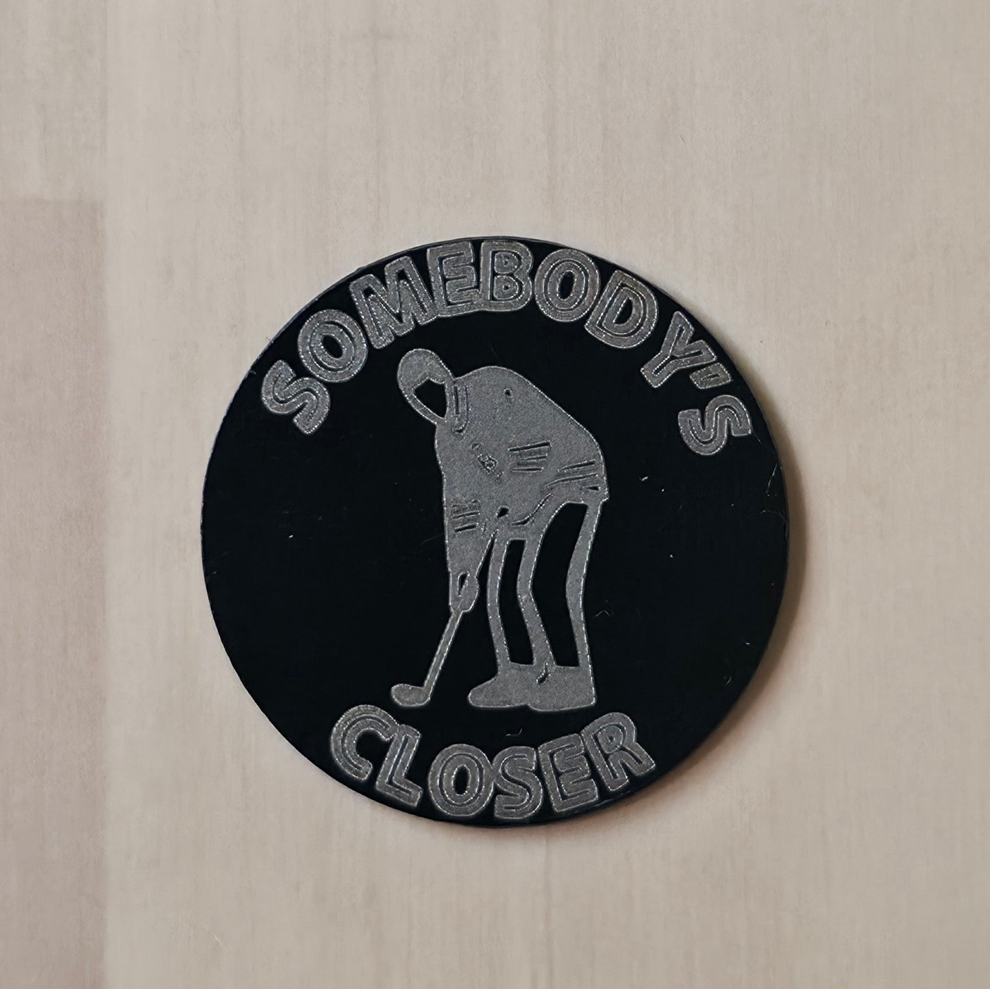 A round black sticker or patch featuring a silver graphic of a person bending forward with a golf club, with the text 'SOMEBODY'S CLOSER' arched above and below, shown on a light wood background.