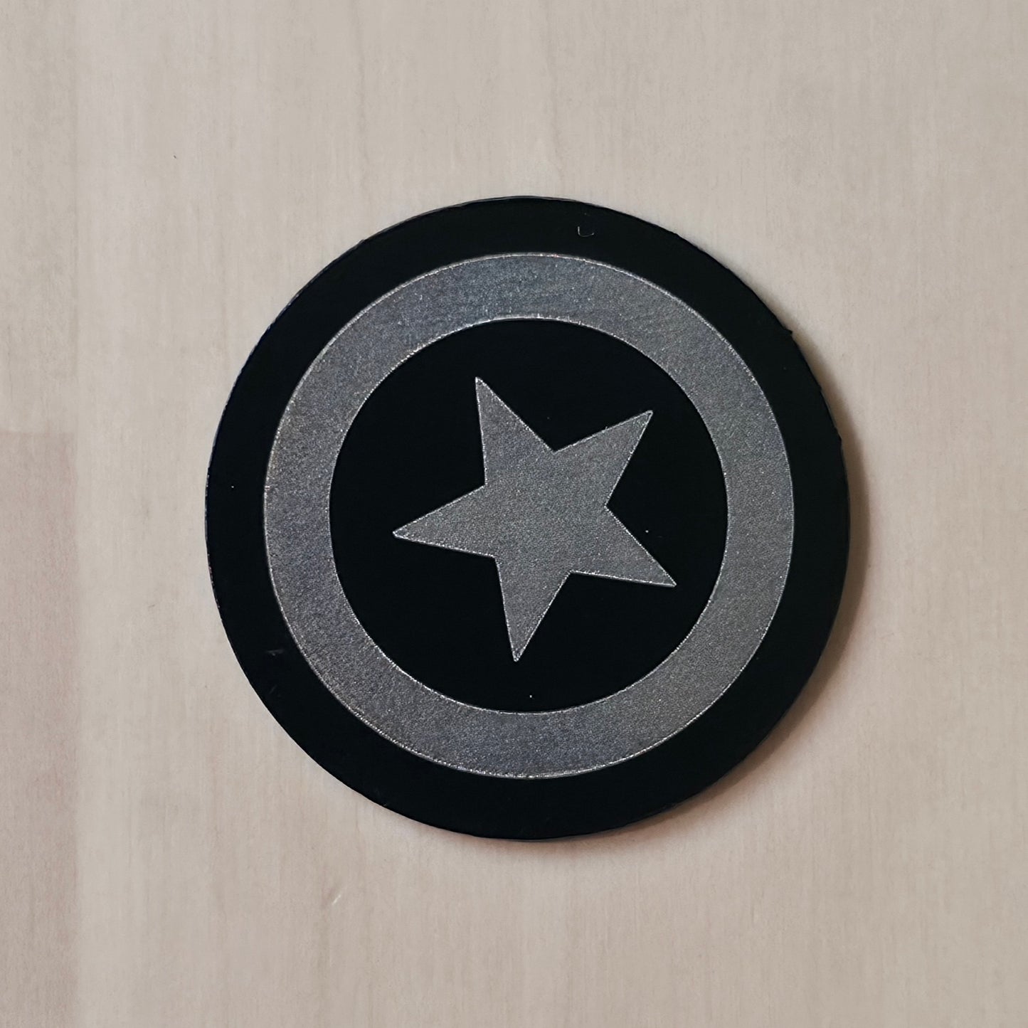 A circular black and silver Captain America shield sticker or decal, shown on a light beige wood-grain surface.