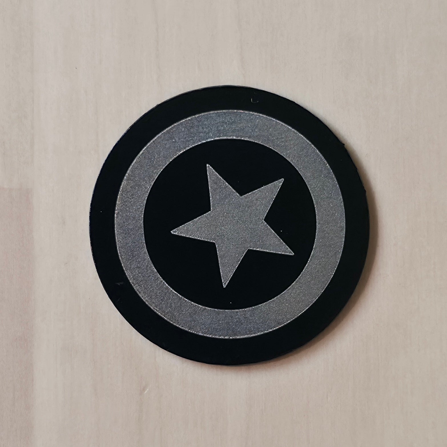 A circular black and silver Captain America shield sticker or decal, shown on a light beige wood-grain surface.