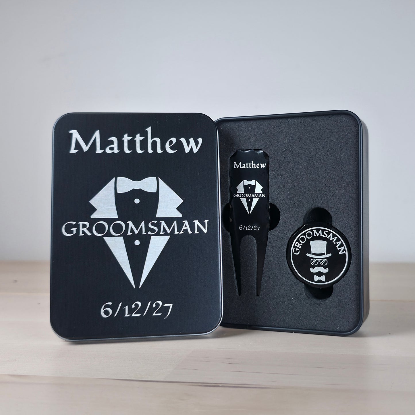 A sleek, personalized black metal golf divot tool and ball marker set rests in a custom engraved tin box. The divot tool displays a tuxedo design,  GROOMSMAN. The round ball marker features a fun top hat, sunglasses, and mustache motif. Both items are securely nestled in a black foam insert. The matching matte black tin cover perfectly mirrors the elegant tuxedo design, creating a memorable, ready-to-gift keepsake for all wedding party members.