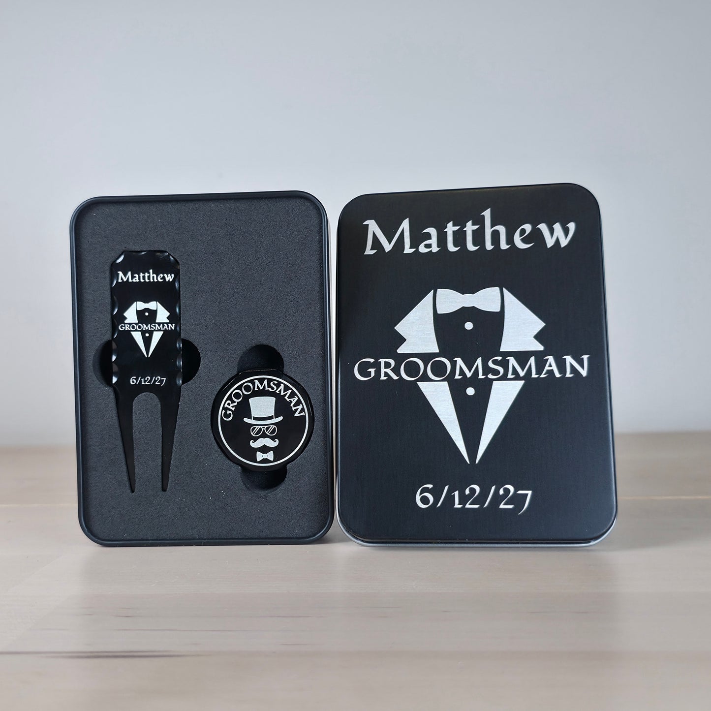 Personalized black metal gift set containing a golf divot tool and a groomsman pin, displayed next to its engraved black tin box on a light wooden surface. A sleek, personalized black metal golf divot tool and ball marker set rests in a custom engraved tin box. The divot tool displays a tuxedo design,  GROOMSMAN. The round ball marker features a fun top hat, sunglasses, and mustache motif. Both items are securely nestled in a black foam insert. The matching matte black tin cover perfectly mirrors the elegan