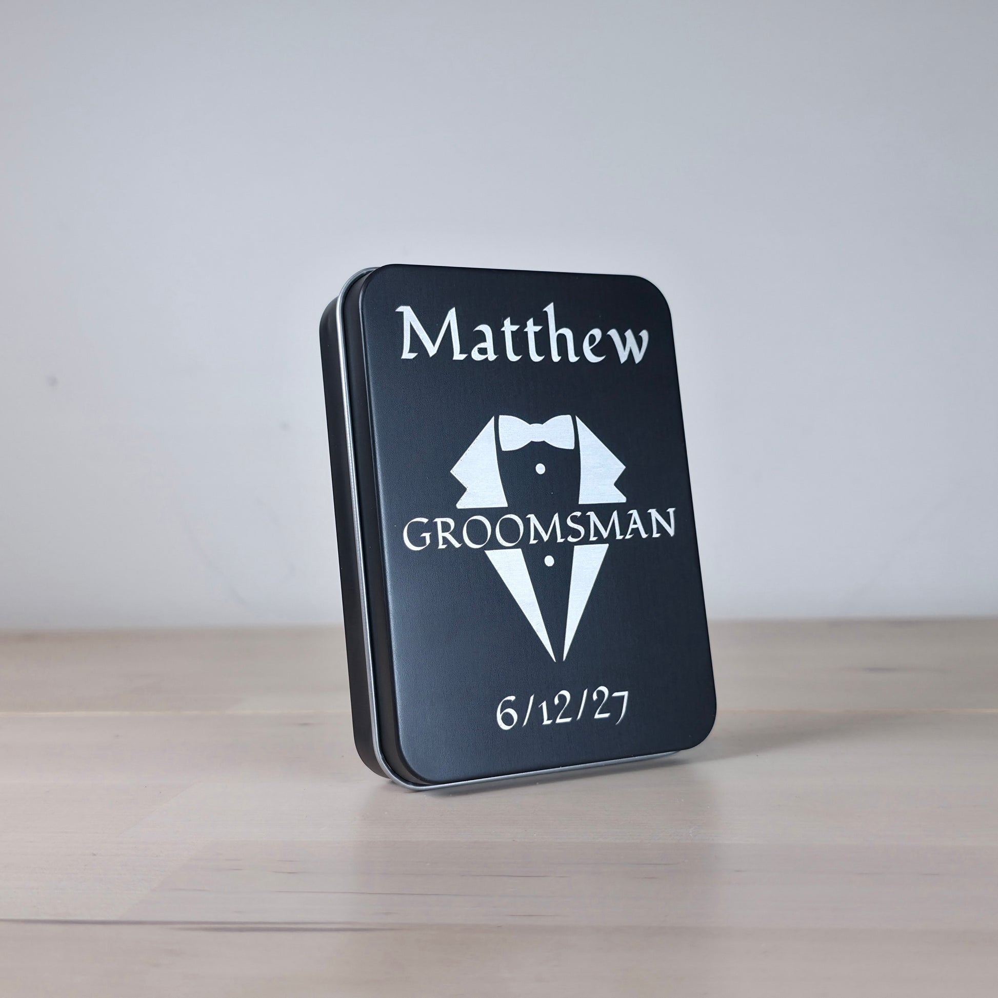 A personalized black metal tin for a groomsman, featuring a white engraved tuxedo graphic and the name 'Matthew' with the date '6/12/27', standing on a light wooden surface against a neutral gray background.