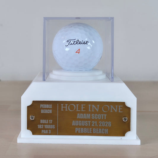 A custom golf trophy display featuring a white Titleist golf ball inside a clear acrylic case, mounted on a white pedestal base with a custom bronze plaque engraved with personalization details for a hole-in-one. This custom golf ball display case rests on artificial green turf. It features a geometric 3D-printed base in matte black or crisp white. A clear acrylic cube sits on top to protect a standard white golf ball. On the front of the base is a rectangular brass metal plaque fastened with two silver scr