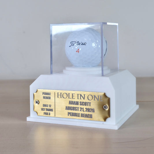 A custom golf trophy base featuring a white pedestal with a gold engraved plaque and a clear acrylic display case holding a white golf ball with a signature. This custom golf ball display case rests on artificial green turf. It features a geometric 3D-printed base in matte black or crisp white. A clear acrylic cube sits on top to protect a standard white golf ball. On the front of the base is a rectangular brass metal plaque fastened with two silver screws. The plaque can be left blank or laser-engraved wit