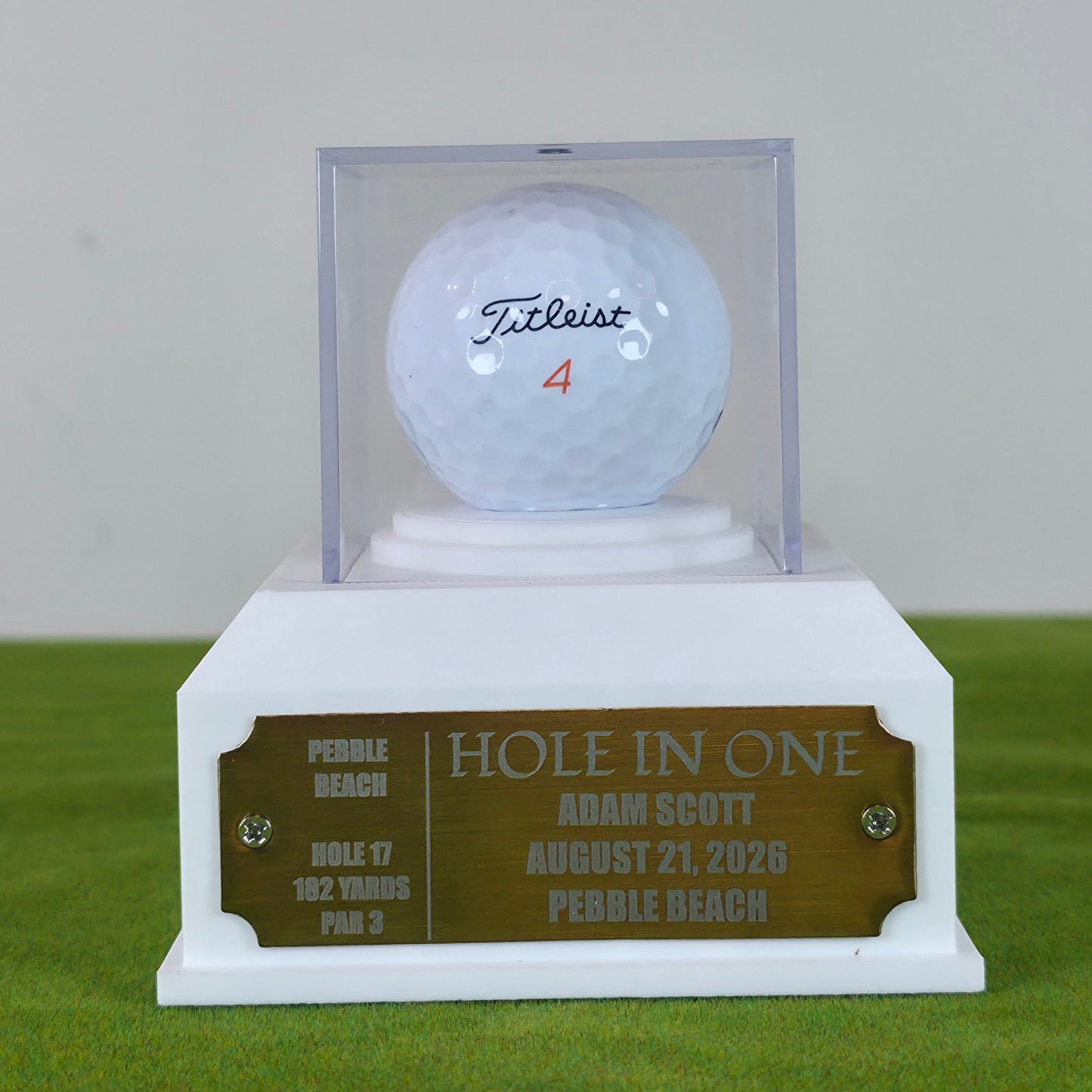 A custom golf trophy featuring a white base with a bronze engraved plaque and a clear acrylic display case holding a white Titleist golf ball with the number 4. This custom golf ball display case rests on artificial green turf. It features a geometric 3D-printed base in matte black or crisp white. A clear acrylic cube sits on top to protect a standard white golf ball. On the front of the base is a rectangular brass metal plaque fastened with two silver screws. The plaque can be left blank or laser-engraved 