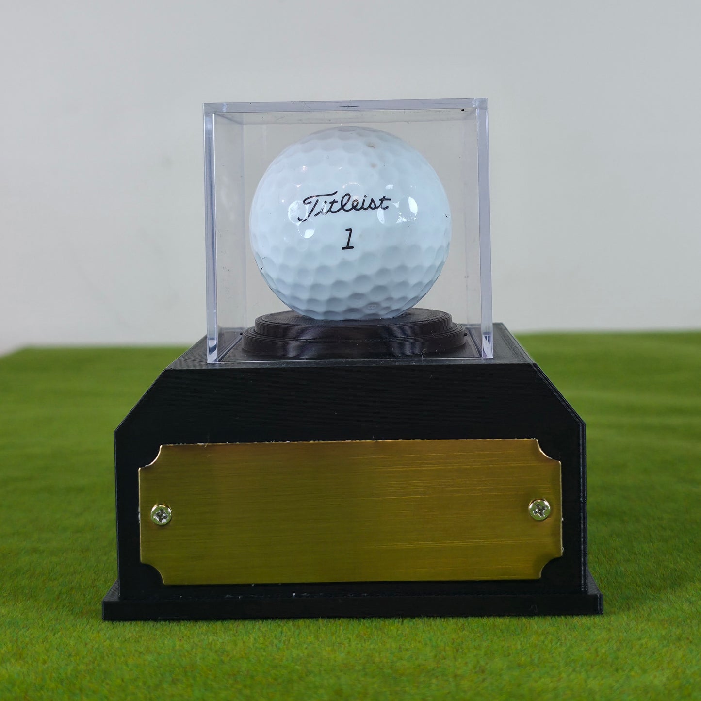 A white Titleist golf ball in a clear acrylic display case, mounted on a black base with a brushed gold front plate, set on artificial green turf against a light gray background. This custom golf ball display case rests on artificial green turf. It features a geometric 3D-printed base in matte black or crisp white. A clear acrylic cube sits on top to protect a standard white golf ball. On the front of the base is a rectangular brass metal plaque fastened with two silver screws. The plaque can be left blank 