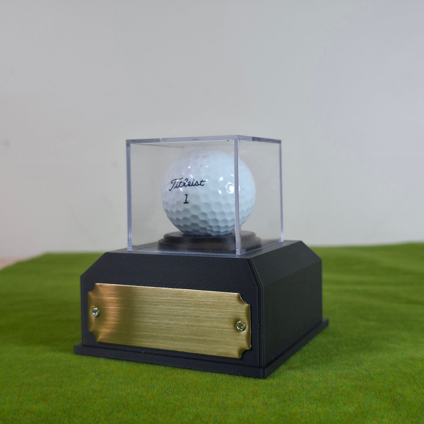 A white Titleist golf ball displayed inside a clear acrylic case on a black base with a gold front plaque, set on a green surface against a light gray background. This custom golf ball display case rests on artificial green turf. It features a geometric 3D-printed base in matte black or crisp white. A clear acrylic cube sits on top to protect a standard white golf ball. On the front of the base is a rectangular brass metal plaque fastened with two silver screws. The plaque can be left blank or laser-engrave
