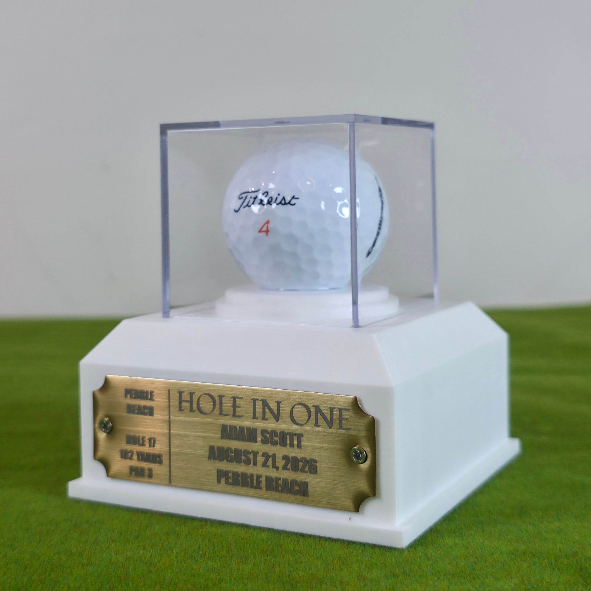 A custom golf trophy display featuring a white golf ball signed by Titleist with the number 4, sealed inside a clear acrylic display case and mounted on a white pedestal base with a gold-colored 'Hole in One' plaque. This custom golf ball display case rests on artificial green turf. It features a geometric 3D-printed base in matte black or crisp white. A clear acrylic cube sits on top to protect a standard white golf ball. On the front of the base is a rectangular brass metal plaque fastened with two silver