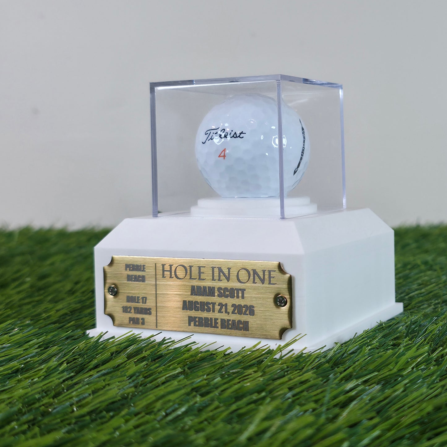 A custom golf trophy display featuring a white base with a gold engraved plaque, a clear acrylic dome, and a white Titleist golf ball on a white stand, set on artificial green turf. This custom golf ball display case rests on artificial green turf. It features a geometric 3D-printed base in matte black or crisp white. A clear acrylic cube sits on top to protect a standard white golf ball. On the front of the base is a rectangular brass metal plaque fastened with two silver screws. The plaque can be left bla