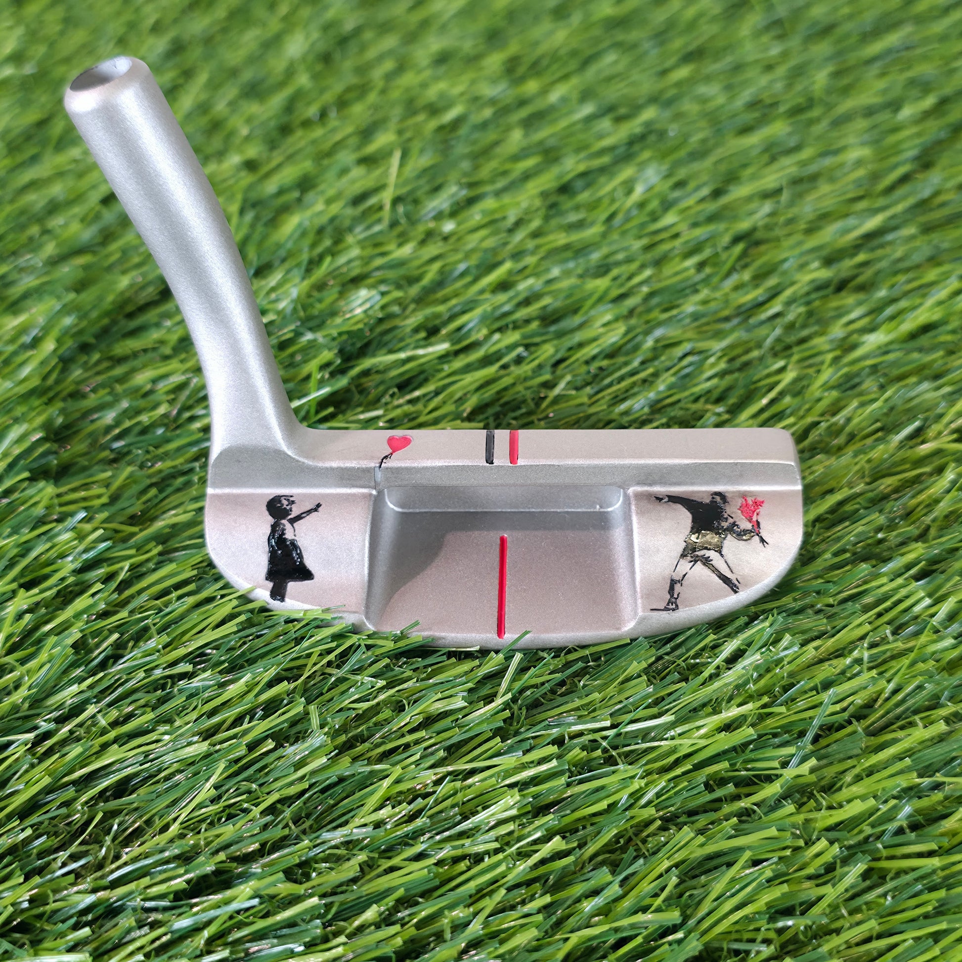 A silver metal golf putter featuring Banksy-style street art prints of a child reaching for a red balloon and a golfer, resting on green artificial turf. Silver golf chipper on green turf. Back features black and red laser-engraved street art silhouettes: a girl reaching for a red heart balloon, and a man throwing red flowers. Top line has a red heart and black/red alignment stripes. The front face shows standard horizontal scoring lines. The metal club head connects to a silver shaft at the heel. These hig