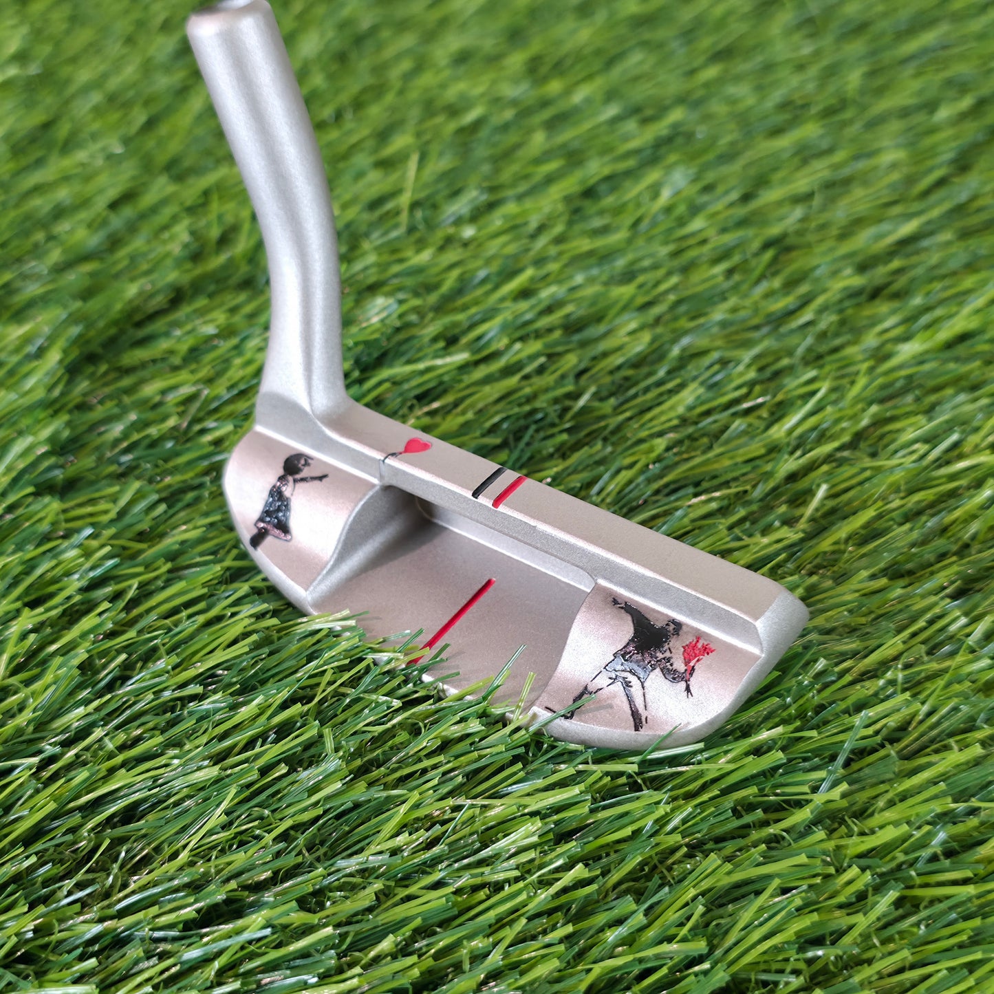 A silver metal golf putter with black and red graphics, resting on green artificial turf. Silver golf chipper on green turf. Back features black and red laser-engraved street art silhouettes: a girl reaching for a red heart balloon, and a man throwing red flowers. Top line has a red heart and black/red alignment stripes. The front face shows standard horizontal scoring lines. The metal club head connects to a silver shaft at the heel. These high-contrast deep engravings are meticulously crafted. Perfect vis