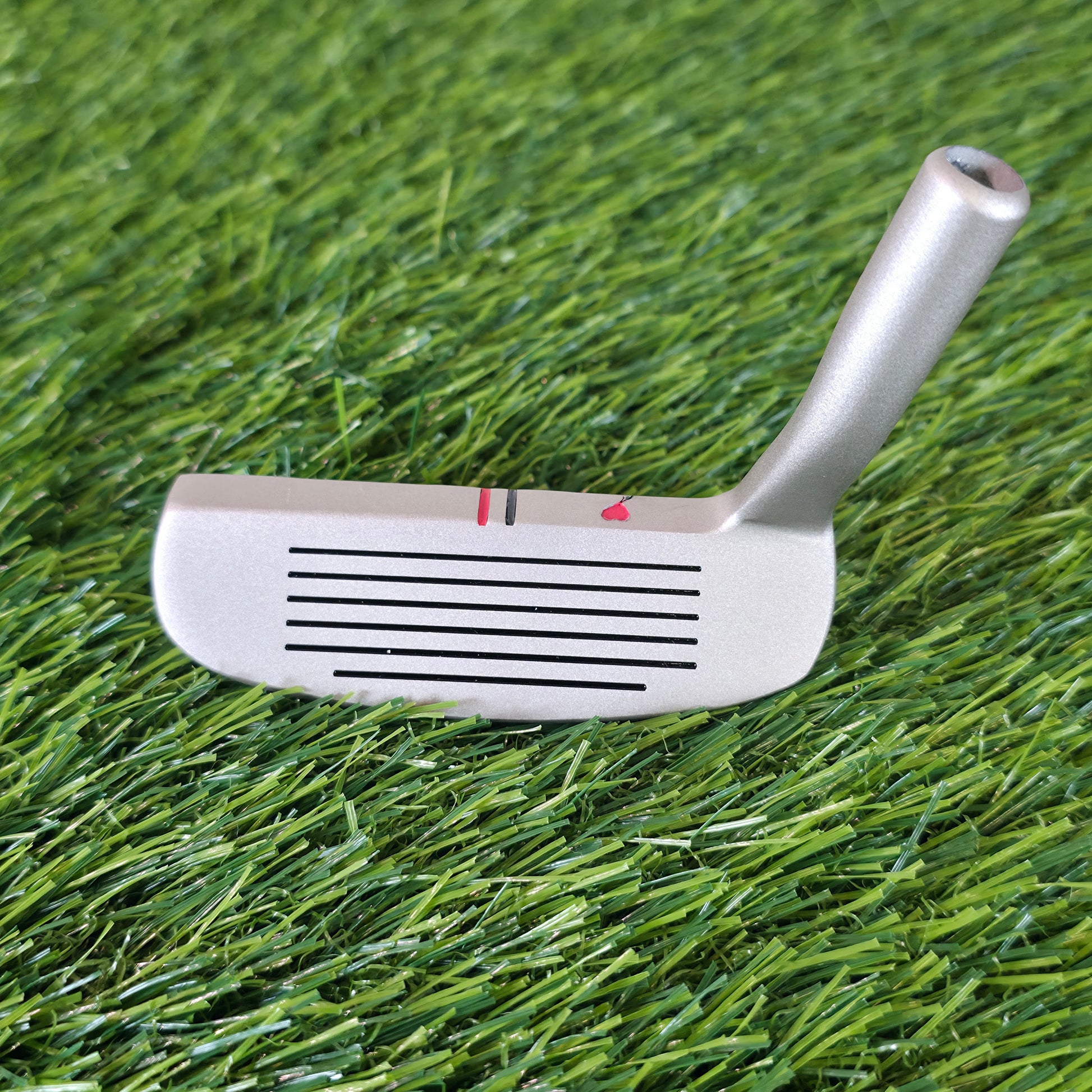 A silver metal golf putter head with black slot lines and small red and black alignment markers, resting on green artificial turf. Silver golf chipper on green turf. Back features black and red laser-engraved street art silhouettes: a girl reaching for a red heart balloon, and a man throwing red flowers. Top line has a red heart and black/red alignment stripes. The front face shows standard horizontal scoring lines. The metal club head connects to a silver shaft at the heel. These high-contrast deep engrave