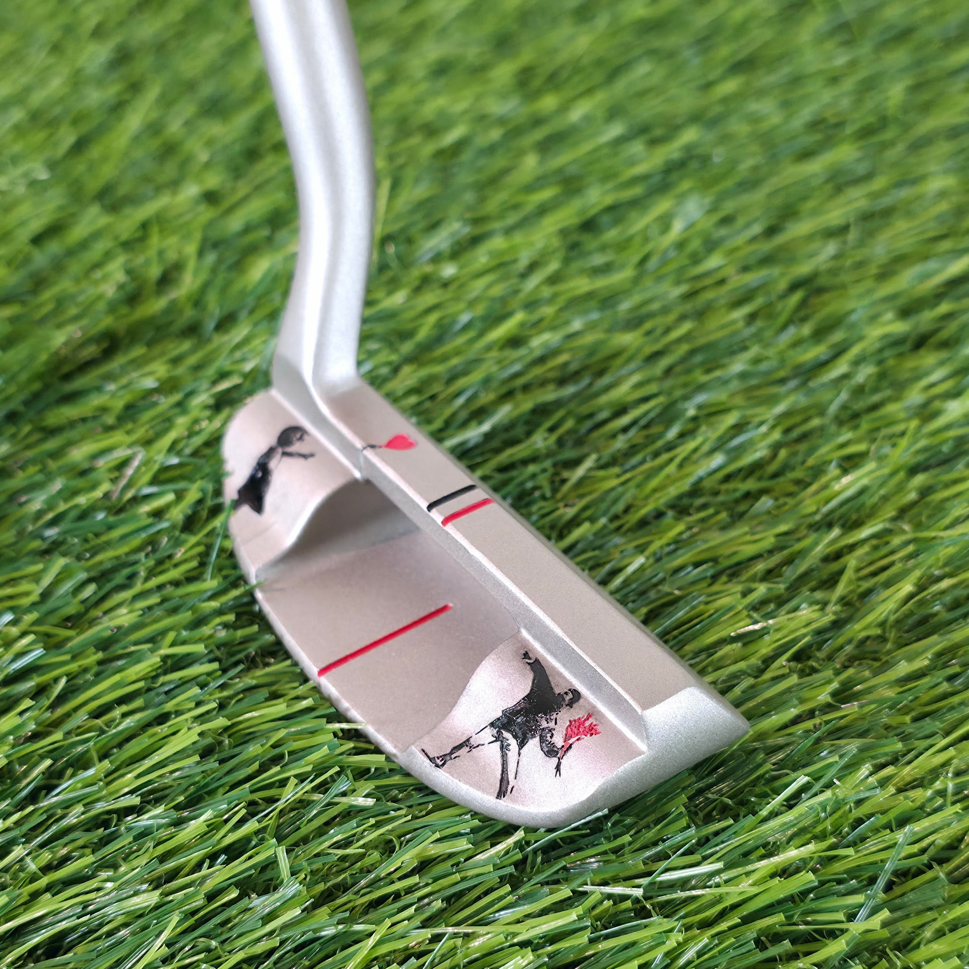 A silver golf putter with black and red graphic designs resting on green artificial turf. Silver golf chipper on green turf. Back features black and red laser-engraved street art silhouettes: a girl reaching for a red heart balloon, and a man throwing red flowers. Top line has a red heart and black/red alignment stripes. The front face shows standard horizontal scoring lines. The metal club head connects to a silver shaft at the heel. These high-contrast deep engravings are meticulously crafted. Perfect vis