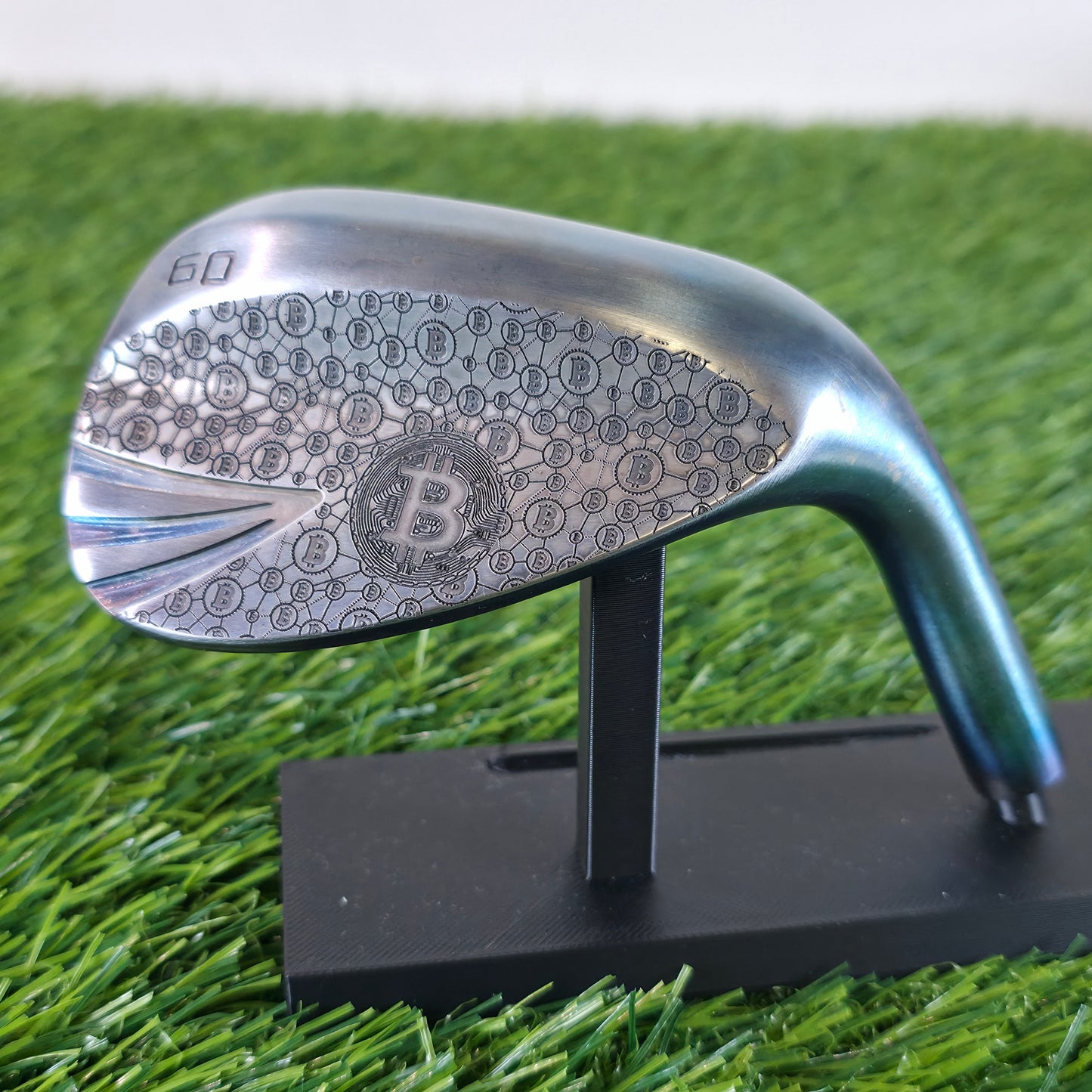 A 60-degree golf lob wedge featuring a custom laser-engraved Bitcoin theme. The back of the metal clubhead displays a central Bitcoin logo surrounded by a complex network of smaller Bitcoin icons and interconnected blockchain nodes. The club face features standard scoring grooves with a detailed engraving of a Bitcoin rocket ship on the toe. Resting on vibrant green artificial turf, this unique wedge seamlessly combines modern cryptocurrency culture with premium custom golf equipment design.