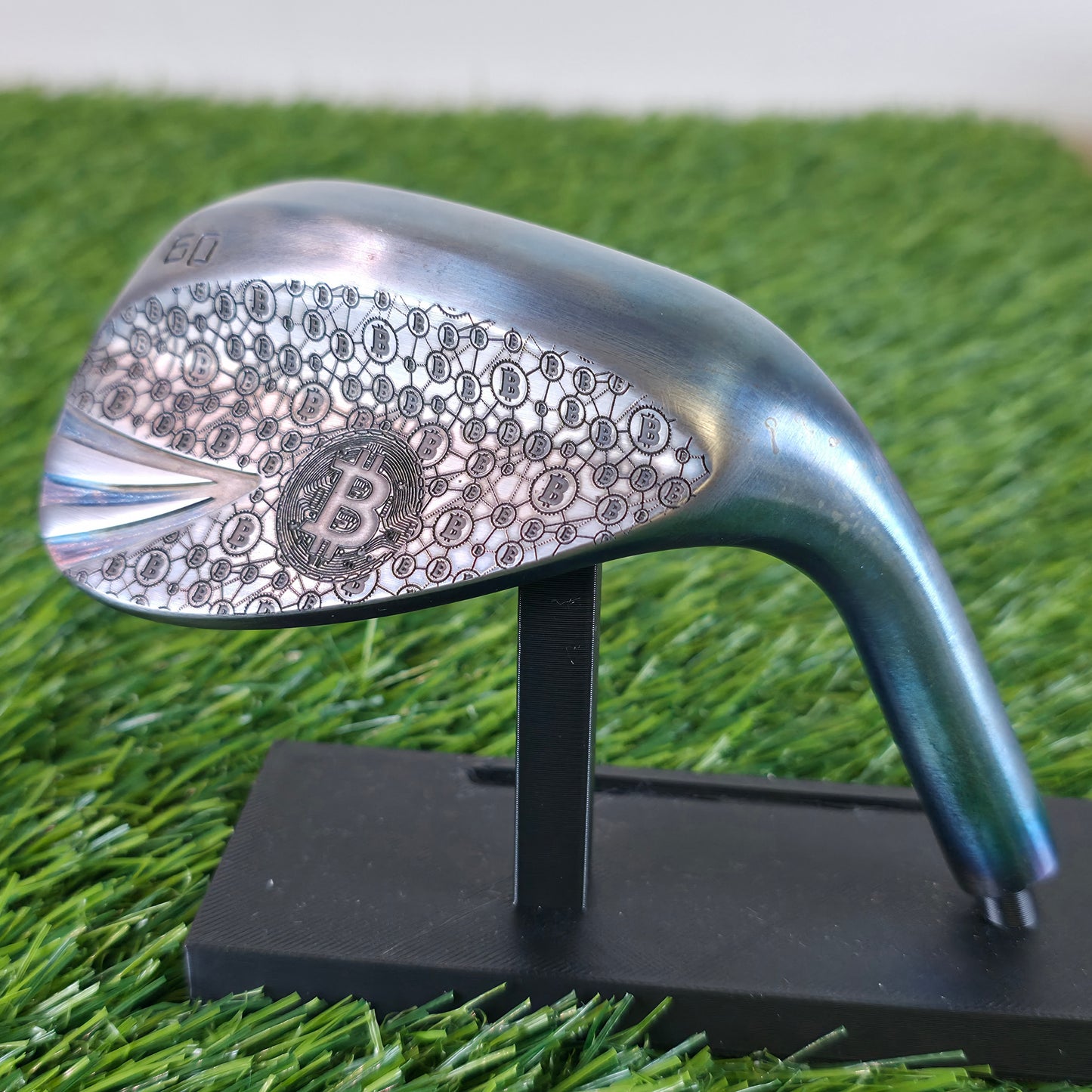 A 60-degree golf lob wedge featuring a custom laser-engraved Bitcoin theme. The back of the metal clubhead displays a central Bitcoin logo surrounded by a complex network of smaller Bitcoin icons and interconnected blockchain nodes. The club face features standard scoring grooves with a detailed engraving of a Bitcoin rocket ship on the toe. Resting on vibrant green artificial turf, this unique wedge seamlessly combines modern cryptocurrency culture with premium custom golf equipment design.