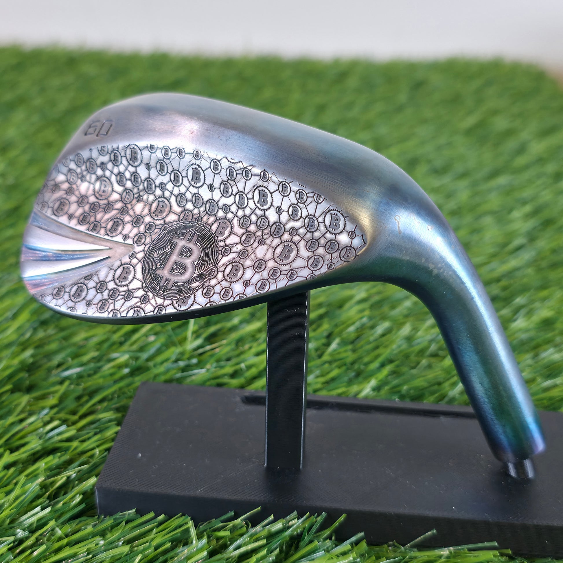 A 60-degree golf lob wedge featuring a custom laser-engraved Bitcoin theme. The back of the metal clubhead displays a central Bitcoin logo surrounded by a complex network of smaller Bitcoin icons and interconnected blockchain nodes. The club face features standard scoring grooves with a detailed engraving of a Bitcoin rocket ship on the toe. Resting on vibrant green artificial turf, this unique wedge seamlessly combines modern cryptocurrency culture with premium custom golf equipment design.