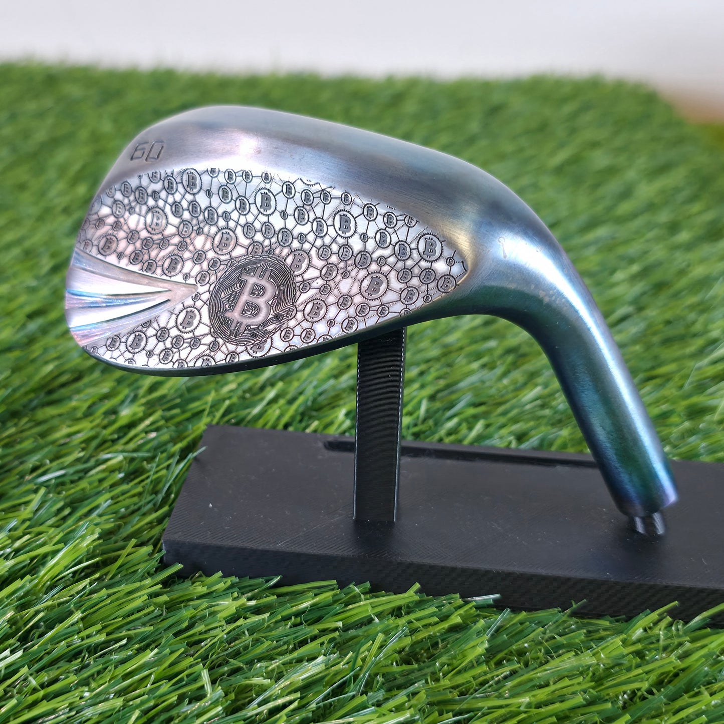 A 60-degree golf lob wedge featuring a custom laser-engraved Bitcoin theme. The back of the metal clubhead displays a central Bitcoin logo surrounded by a complex network of smaller Bitcoin icons and interconnected blockchain nodes. The club face features standard scoring grooves with a detailed engraving of a Bitcoin rocket ship on the toe. Resting on vibrant green artificial turf, this unique wedge seamlessly combines modern cryptocurrency culture with premium custom golf equipment design.
