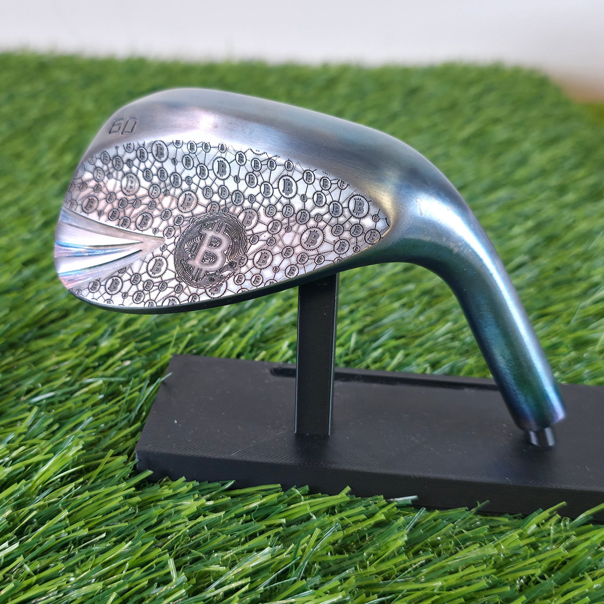 A 60-degree golf lob wedge featuring a custom laser-engraved Bitcoin theme. The back of the metal clubhead displays a central Bitcoin logo surrounded by a complex network of smaller Bitcoin icons and interconnected blockchain nodes. The club face features standard scoring grooves with a detailed engraving of a Bitcoin rocket ship on the toe. Resting on vibrant green artificial turf, this unique wedge seamlessly combines modern cryptocurrency culture with premium custom golf equipment design.