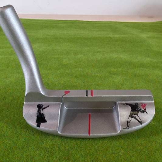 A silver metallic golf putter featuring Banksy-style street art illustrations of a child and an adult holding a red balloon, displayed on a green artificial turf background. Silver golf chipper on green turf. Back features black and red laser-engraved street art silhouettes: a girl reaching for a red heart balloon, and a man throwing red flowers. Top line has a red heart and black/red alignment stripes. The front face shows standard horizontal scoring lines. The metal club head connects to a silver shaft at