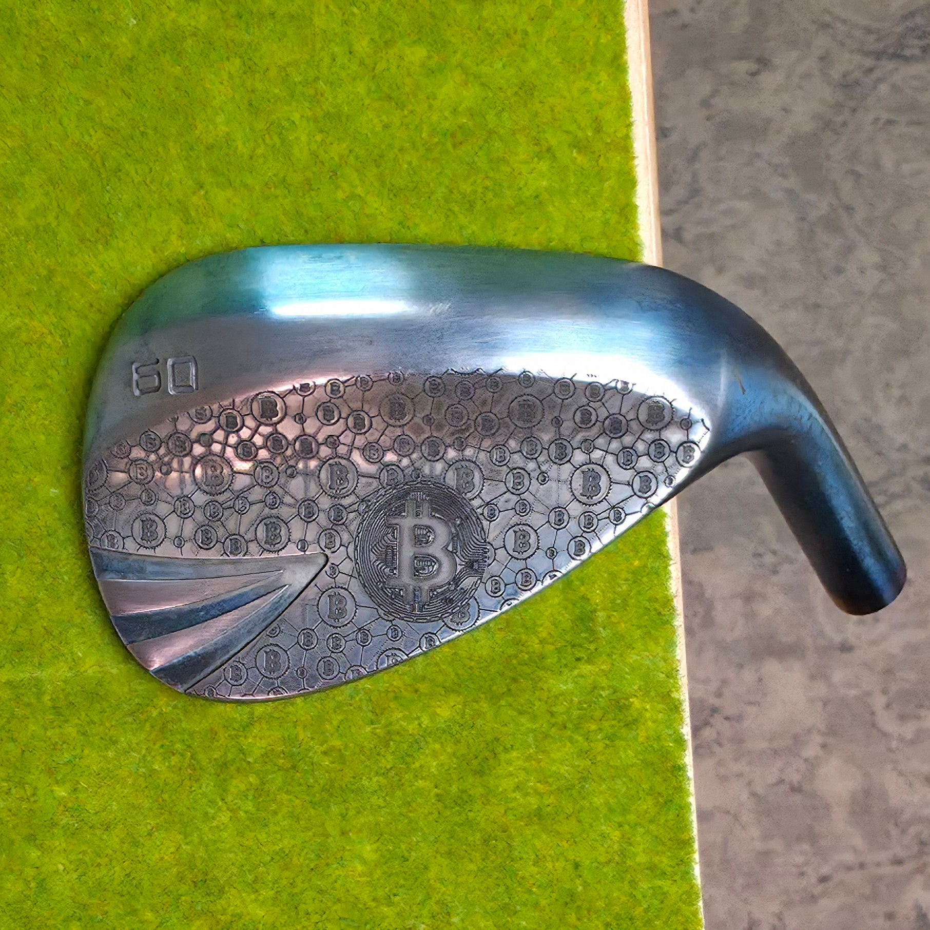 A 60-degree golf lob wedge featuring a custom laser-engraved Bitcoin theme. The back of the metal clubhead displays a central Bitcoin logo surrounded by a complex network of smaller Bitcoin icons and interconnected blockchain nodes. The club face features standard scoring grooves with a detailed engraving of a Bitcoin rocket ship on the toe. Resting on vibrant green artificial turf, this unique wedge seamlessly combines modern cryptocurrency culture with premium custom golf equipment design.