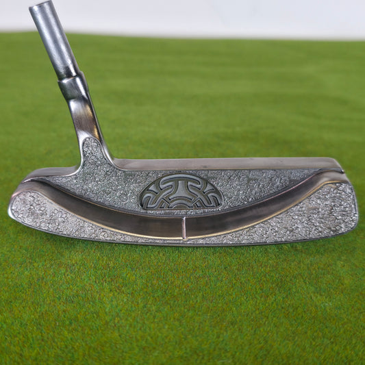 A close-up of a silver metal golf putter with a textured finish and an embossed circular logo, resting on a green artificial turf background. A detailed close-up of a custom silver blade golf putter resting on green turf. The golf club features a classic plumber's neck hosel and a smooth polished top line. The back cavity and bumpers showcase a deep, rugged stippled laser-engraved texture that dramatically contrasts with the sleek top. Inside the central cavity sits a raised, semi-circular stylized geometri