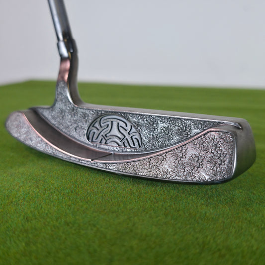 A detailed close-up of a custom silver blade golf putter resting on green turf. The golf club features a classic plumber's neck hosel and a smooth polished top line. The back cavity and bumpers showcase a deep, rugged stippled laser-engraved texture that dramatically contrasts with the sleek top. Inside the central cavity sits a raised, semi-circular stylized geometric Celtic knot pattern. the meticulous craftsmanship and the premium metallic finish of this custom club. azalea design pattern