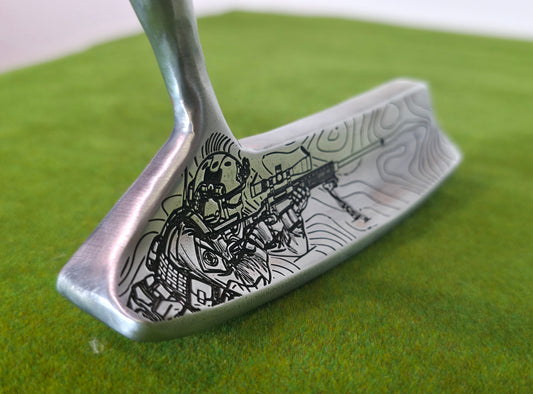 A close-up of a silver metal golf putter with an intricate black line-art illustration of a skeleton holding a rifle, resting on a green artificial turf background.A silver Dunlop blade golf putter on green turf. The sole features a detailed, custom black laser engraving of a tactical military sniper aiming a scoped rifle on a bipod, set against winding topographical map lines. The back cavity has a black section with white Dunlop text and a target logo. The front club face includes a rectangular red insert