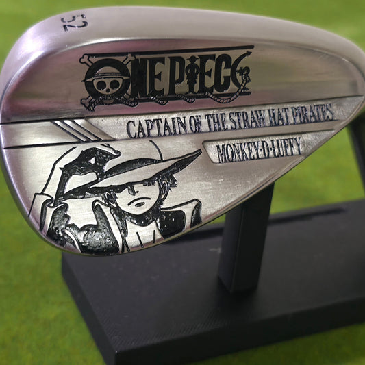 Close-up of a custom-engraved silvery 52-degree golf wedge on green grass. The club head features a deep black laser engraving of Anime Pirate tipping his straw hat.  The Pirate skull logo is etched near the top edge. This brushed steel club blends anime fandom with golf, showcasing high-contrast, permanent artwork. Perfect for display or play, it highlights crisp lines upon a brilliant metallic golf surface.