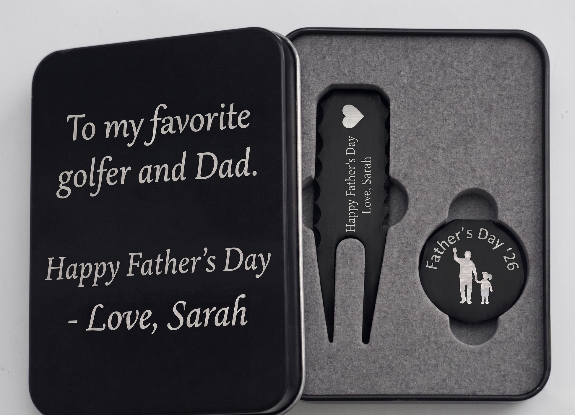 Black metal golf gift set for Father's Day in a custom engraved tin box. Inside sits a customized divot repair tool and a round ball marker resting in foam. Engraving options include sentimental quotes from kids, funny dad golf stats like three-putts, topographic patterns with initials, or classic crossed golf clubs. A perfect personalized fathers or grandpa gift for the golf course. The dark matte metal provides high contrast for the silver laser engraving, making the text pop nicely.