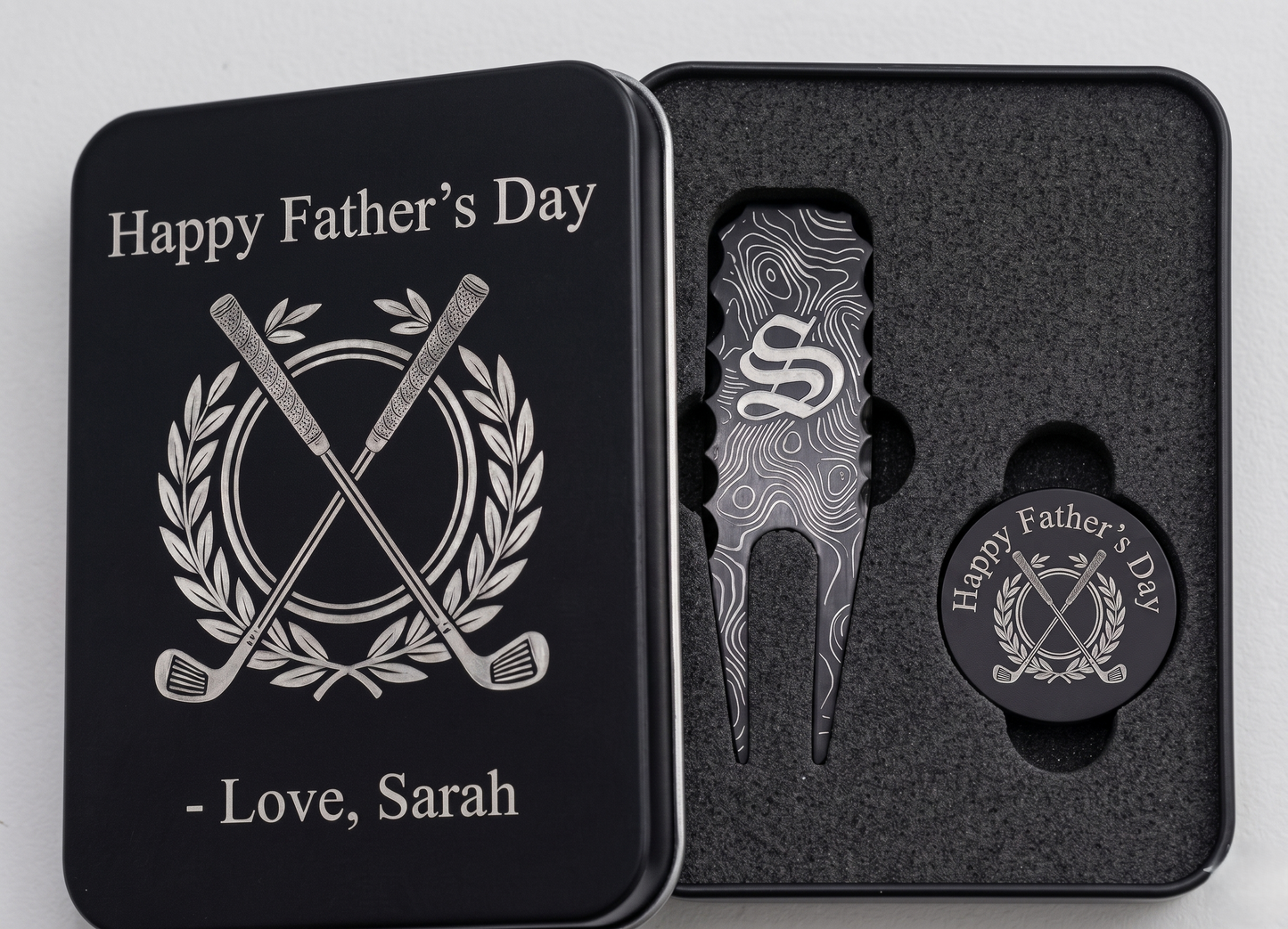 Black metal golf gift set for Father's Day in a custom engraved tin box. Inside sits a customized divot repair tool and a round ball marker resting in foam. Engraving options include sentimental quotes from kids, funny dad golf stats like three-putts, topographic patterns with initials, or classic crossed golf clubs. A perfect personalized fathers or grandpa gift for the golf course. The dark matte metal provides high contrast for the silver laser engraving, making the text pop nicely.