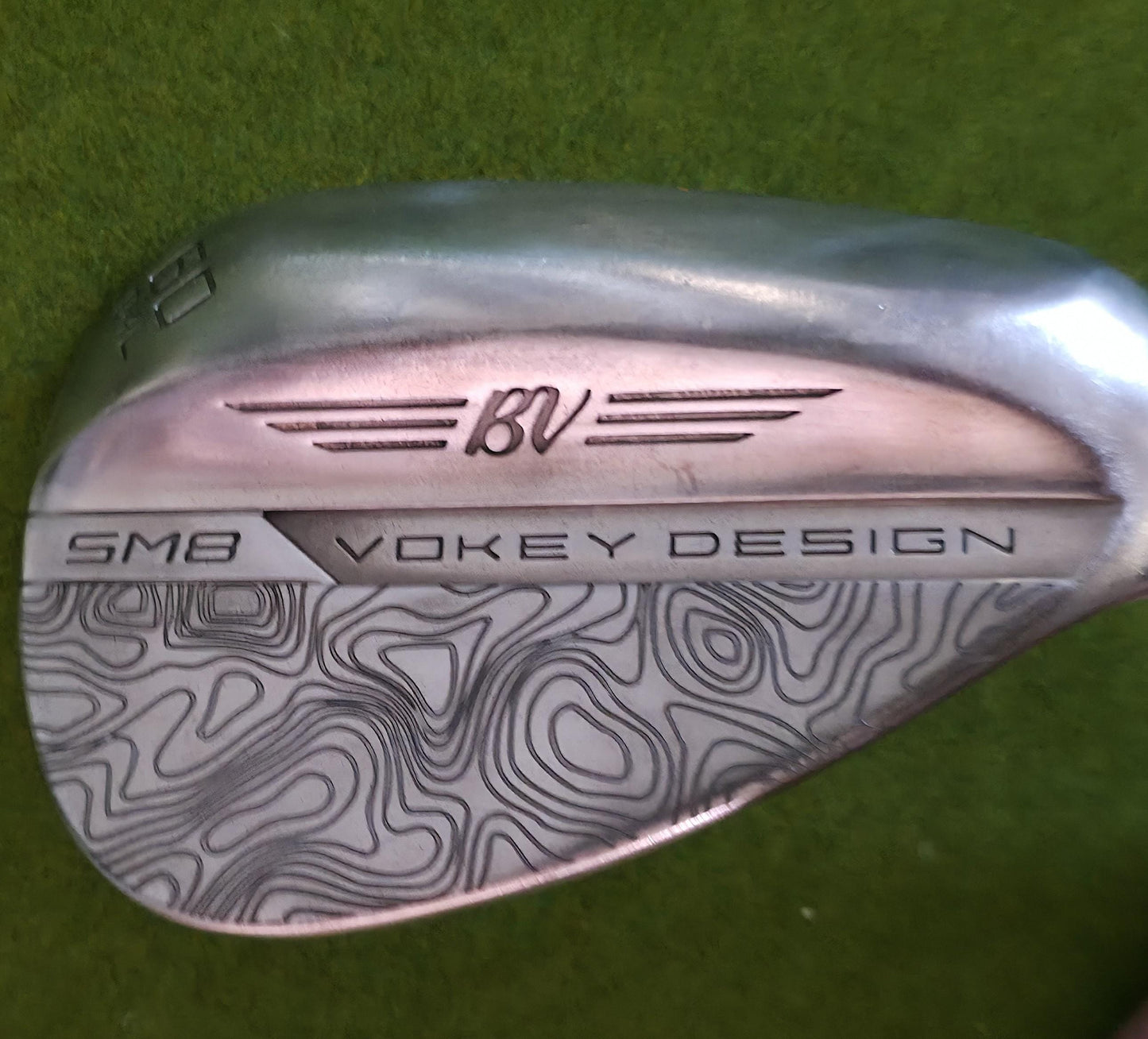 Custom laser-engraved Titleist Vokey SM8 golf wedge by Par Bros Golf, featuring a detailed, wrap-around topographic map pattern. The intricate artisan etching covers the steel clubhead, highlighting unique personalized craftsmanship. Titleist Vokey Design SM8 wedge with deep, intricate topographic map-style laser engraving covering the clubhead's surface. The 'BV' logo, 'SM8', and 'VOKEY DESIGN' markings are clearly visible on the brushed steel finish.