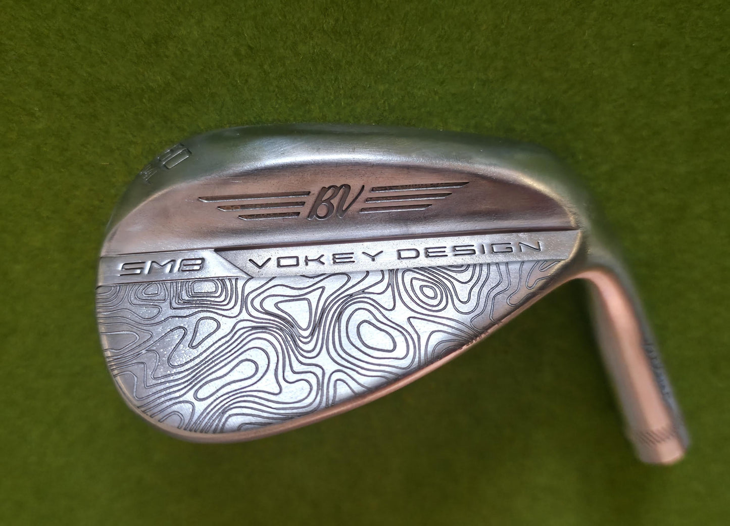 Custom laser-engraved Titleist Vokey SM8 golf wedge by Par Bros Golf, featuring a detailed, wrap-around topographic map pattern. The intricate artisan etching covers the steel clubhead, highlighting unique personalized craftsmanship. Titleist Vokey Design SM8 wedge with deep, intricate topographic map-style laser engraving covering the clubhead's surface. The 'BV' logo, 'SM8', and 'VOKEY DESIGN' markings are clearly visible on the brushed steel finish.