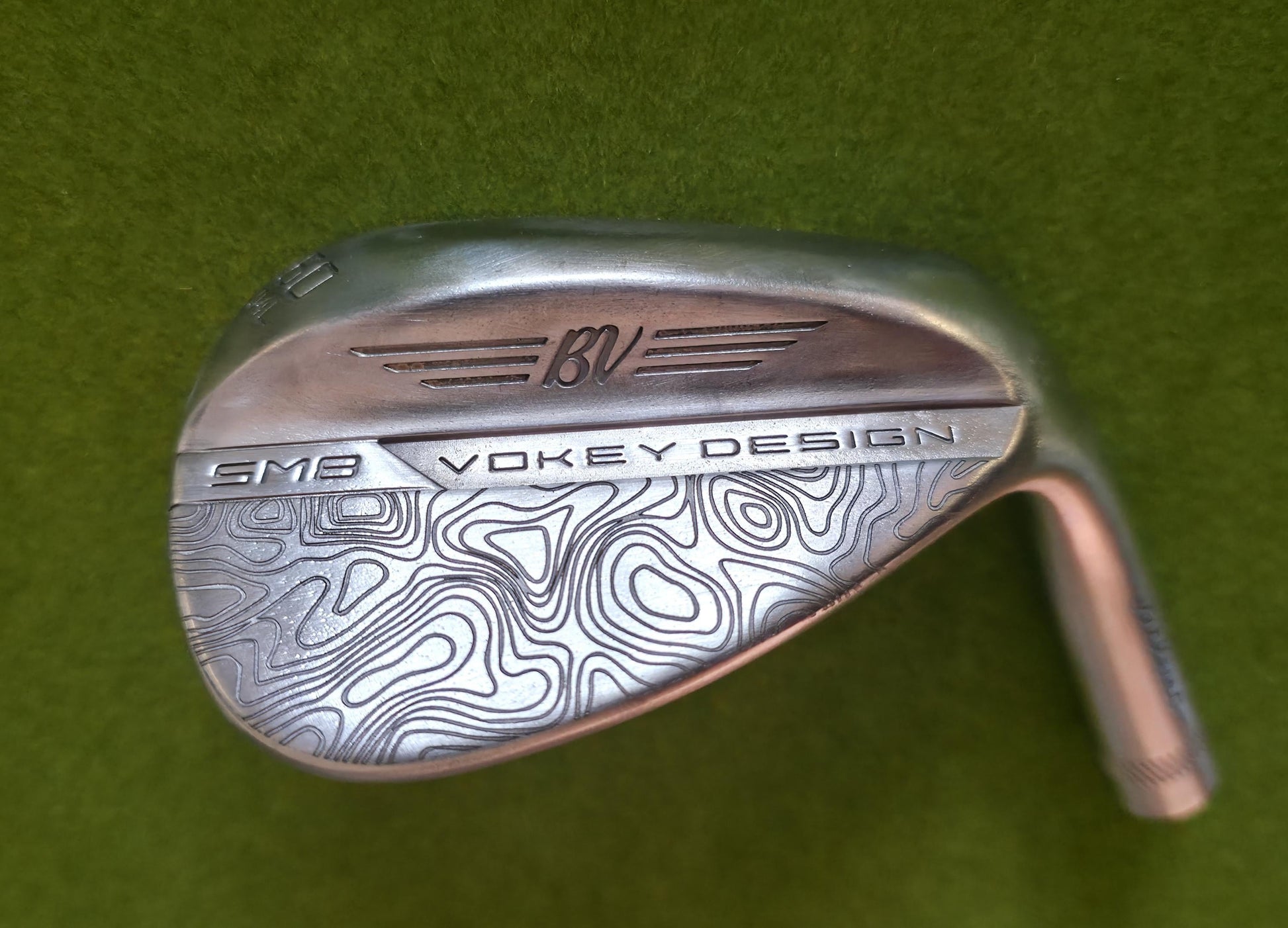 Custom laser-engraved Titleist Vokey SM8 golf wedge by Par Bros Golf, featuring a detailed, wrap-around topographic map pattern. The intricate artisan etching covers the steel clubhead, highlighting unique personalized craftsmanship. Titleist Vokey Design SM8 wedge with deep, intricate topographic map-style laser engraving covering the clubhead's surface. The 'BV' logo, 'SM8', and 'VOKEY DESIGN' markings are clearly visible on the brushed steel finish.