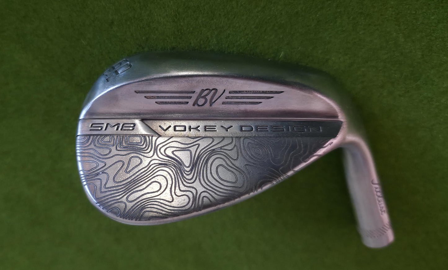 Custom laser-engraved Titleist Vokey SM8 golf wedge by Par Bros Golf, featuring a detailed, wrap-around topographic map pattern. The intricate artisan etching covers the steel clubhead, highlighting unique personalized craftsmanship. Titleist Vokey Design SM8 wedge with deep, intricate topographic map-style laser engraving covering the clubhead's surface. The 'BV' logo, 'SM8', and 'VOKEY DESIGN' markings are clearly visible on the brushed steel finish.