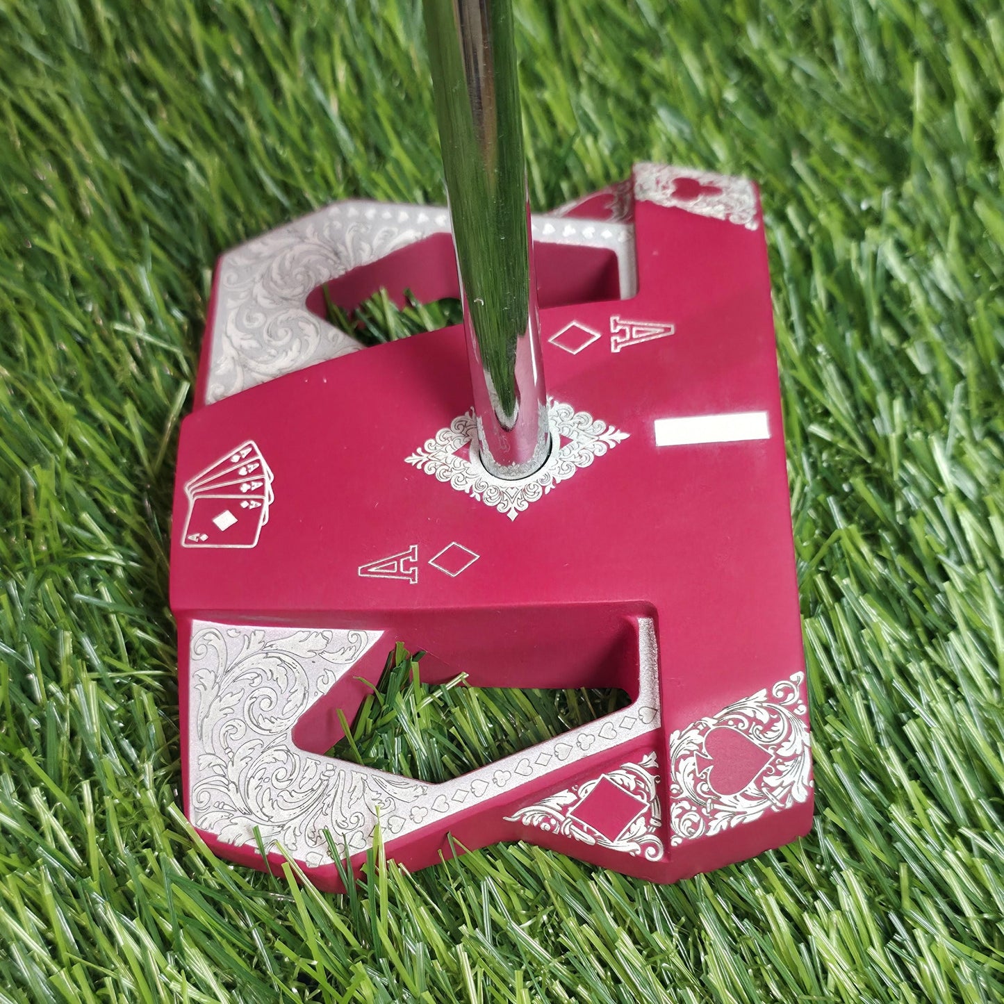 Detailed front view of a custom milled, deep matte red mallet putter head. Intricate playing card suit (Hearts, Diamonds, Spades, Clubs) engravings in satin silver. Features a fanned card graphic, "Ace of Diamonds" symbols, central hole, and center alignment aid. Engraved hearts on corners. ngled top-down view of a custom red mallet putter head resting on green artificial turf, showing the detailed silver scrollwork wings with small suit symbols.