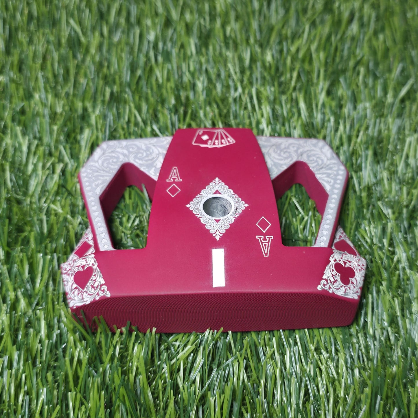 Detailed front view of a custom milled, deep matte red mallet putter head. Intricate playing card suit (Hearts, Diamonds, Spades, Clubs) engravings in satin silver. Features a fanned card graphic, "Ace of Diamonds" symbols, central hole, and center alignment aid. Engraved hearts on corners. angled top-down view of a custom red mallet putter head resting on green artificial turf, showing the detailed silver scrollwork wings with small suit symbols.