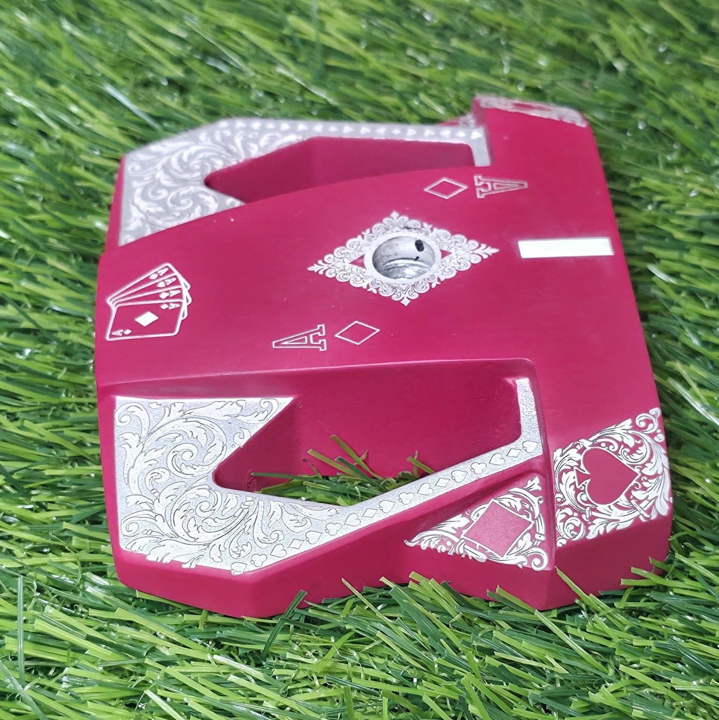Detailed front view of a custom milled, deep matte red mallet putter head. Intricate playing card suit (Hearts, Diamonds, Spades, Clubs) engravings in satin silver. Features a fanned card graphic, "Ace of Diamonds" symbols, central hole, and center alignment aid. Engraved hearts on corners. angled top-down view of a custom red mallet putter head resting on green artificial turf, showing the detailed silver scrollwork wings with small suit symbols.