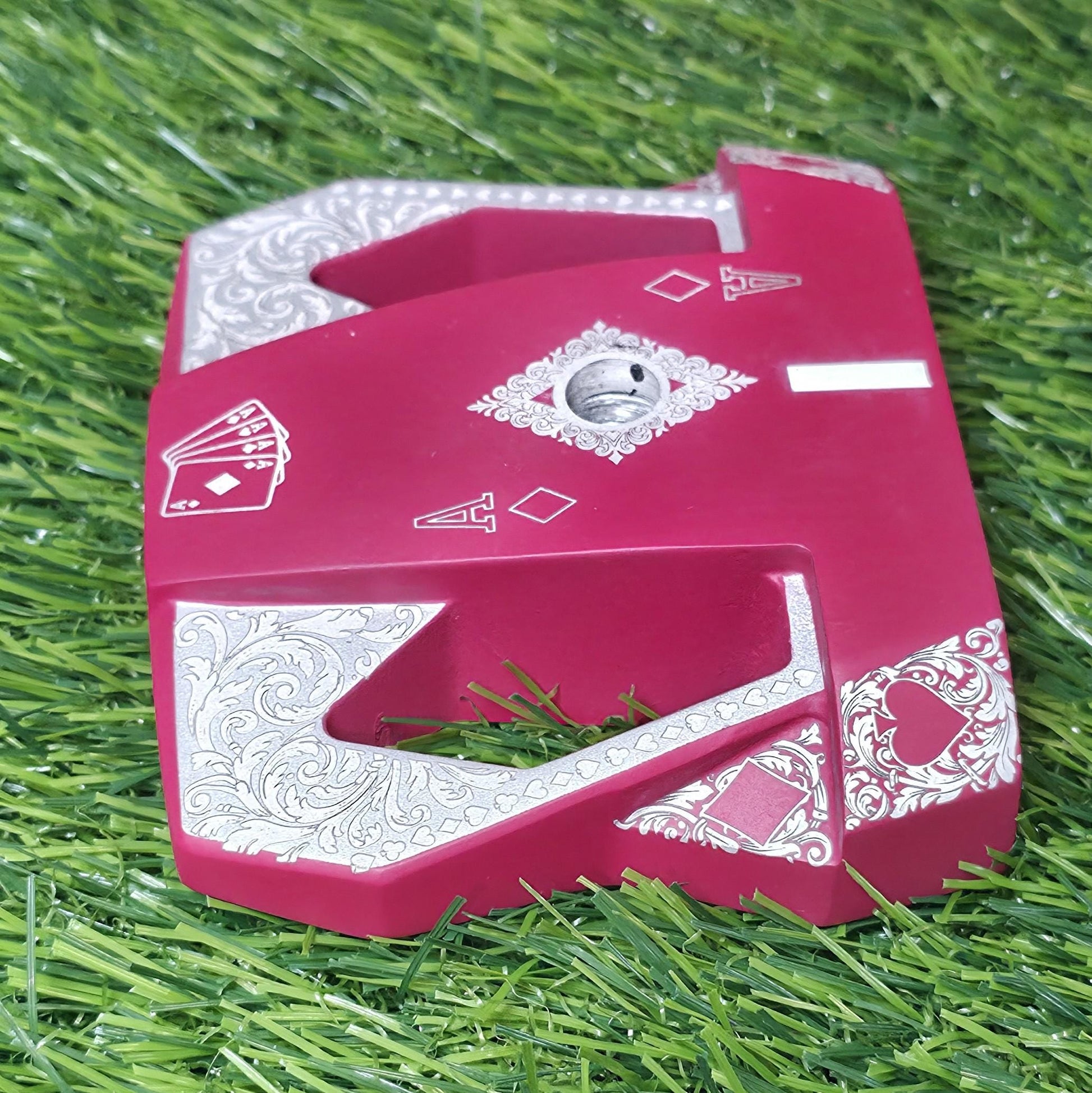 Detailed front view of a custom milled, deep matte red mallet putter head. Intricate playing card suit (Hearts, Diamonds, Spades, Clubs) engravings in satin silver. Features a fanned card graphic, "Ace of Diamonds" symbols, central hole, and center alignment aid. Engraved hearts on corners. angled top-down view of a custom red mallet putter head resting on green artificial turf, showing the detailed silver scrollwork wings with small suit symbols.