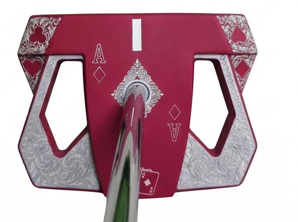 Detailed front view of a custom milled, deep matte red mallet putter head. Intricate playing card suit (Hearts, Diamonds, Spades, Clubs) engravings in satin silver. Features a fanned card graphic, "Ace of Diamonds" symbols, central hole, and center alignment aid. Engraved hearts on corners. ngled top-down view of a custom red mallet putter head resting on green artificial turf, showing the detailed silver scrollwork wings with small suit symbols.