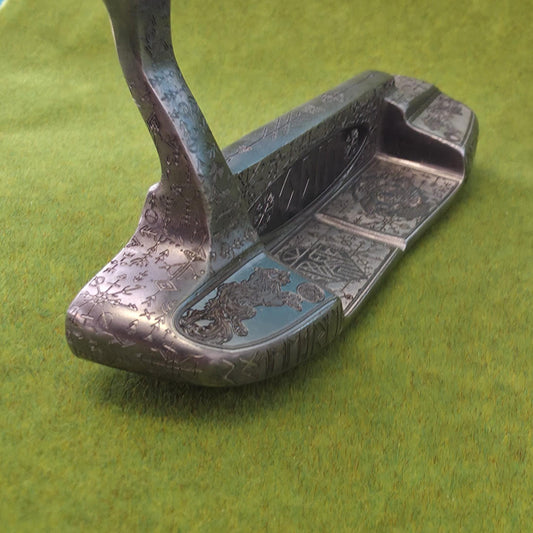 May include: A detailed, antique-style golf putter with intricate engravings. The putter is primarily silver with a dark, textured finish and a curved shaft. The face of the putter features a detailed design, and the putter is set against a green background.