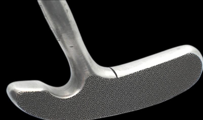 May include: A silver-colored golf club putter with a patterned grip. The putter is angled slightly, showcasing its design and the texture of the grip. The club is set against a marble-like surface.