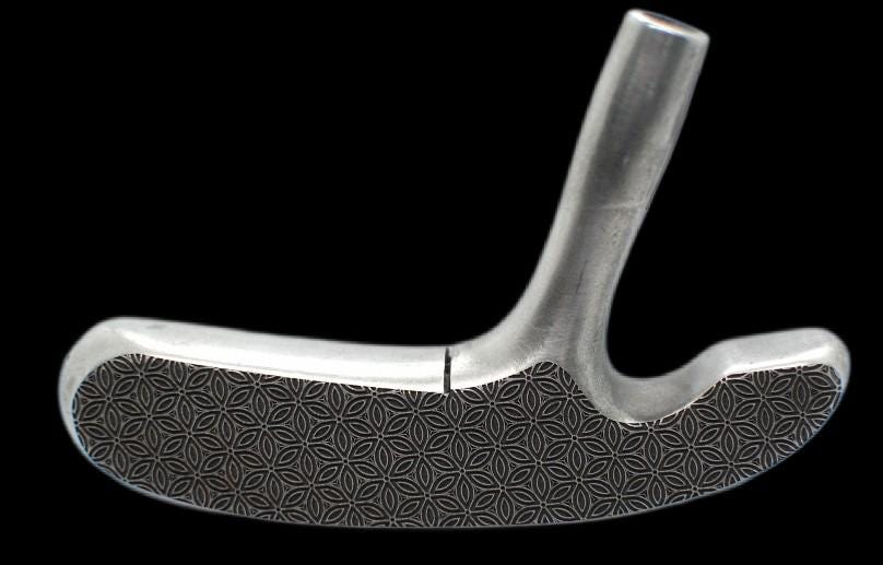 May include: A silver-colored golf club putter with a patterned grip. The putter is angled slightly, showcasing its design and the texture of the grip. The club is set against a marble-like surface.